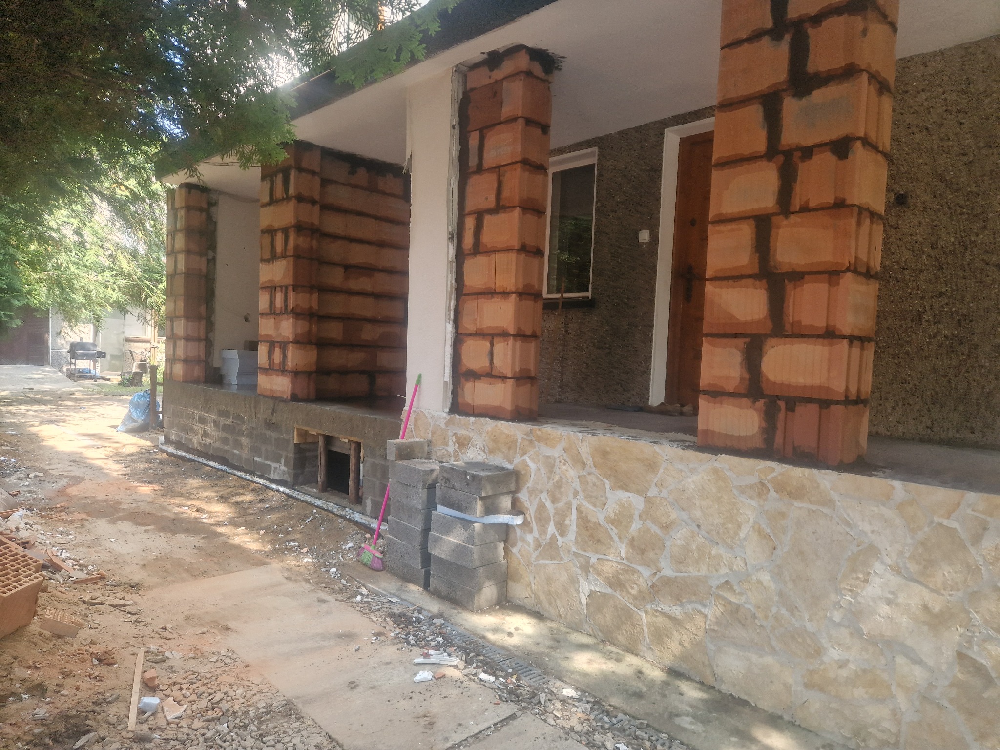 Budowa ganku domu jednorodzinnego z kolumnami z czerwonej cegły, widoczne bloczki betonowe i fragment elewacji z kamienia naturalnego.