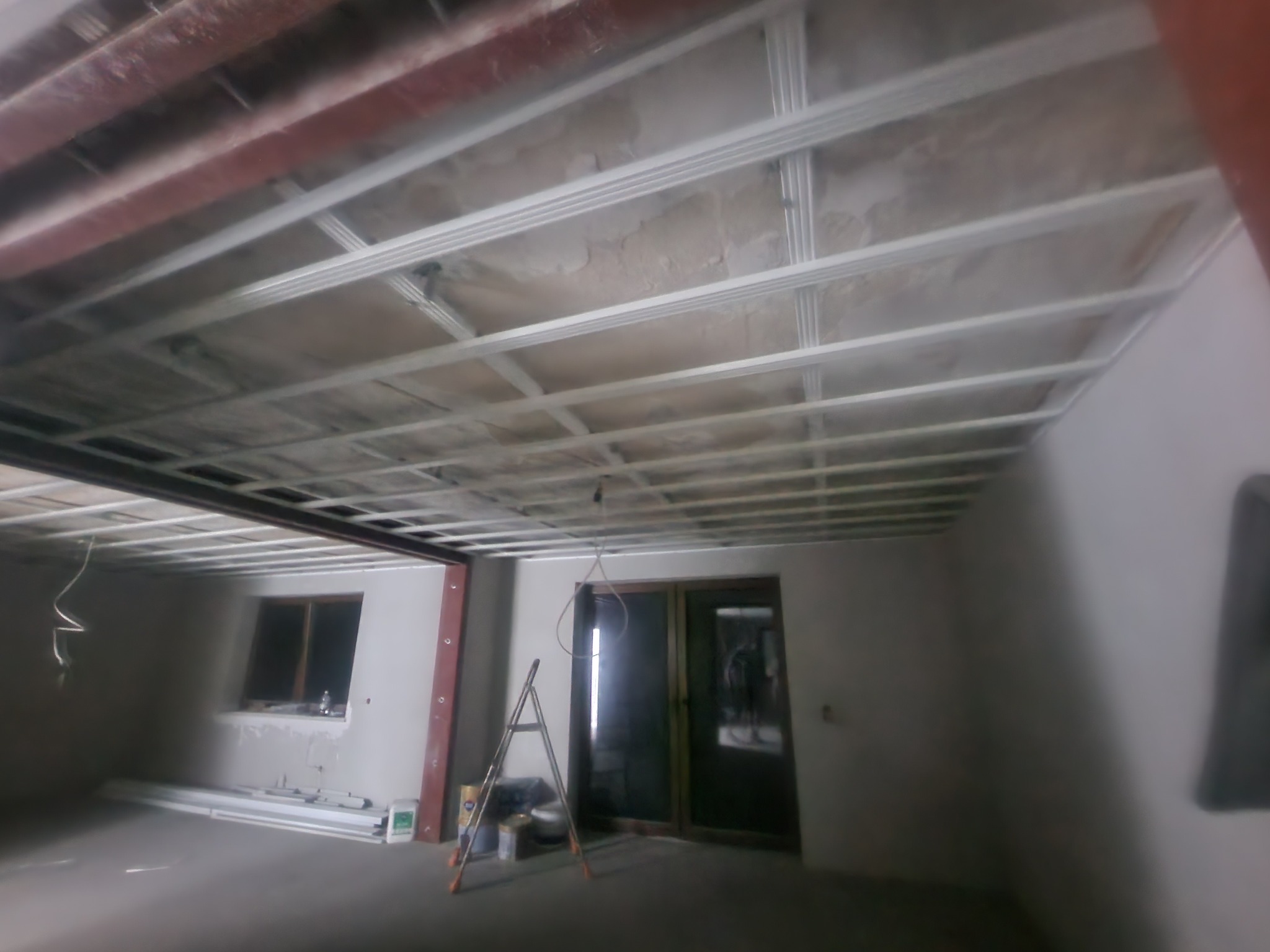 Wnętrze w trakcie prac wykończeniowych: widoczny stelaż sufitu podwieszanego, drabina aluminiowa i materiały budowlane przy ścianie z oknem i drzwiami balkonowymi.