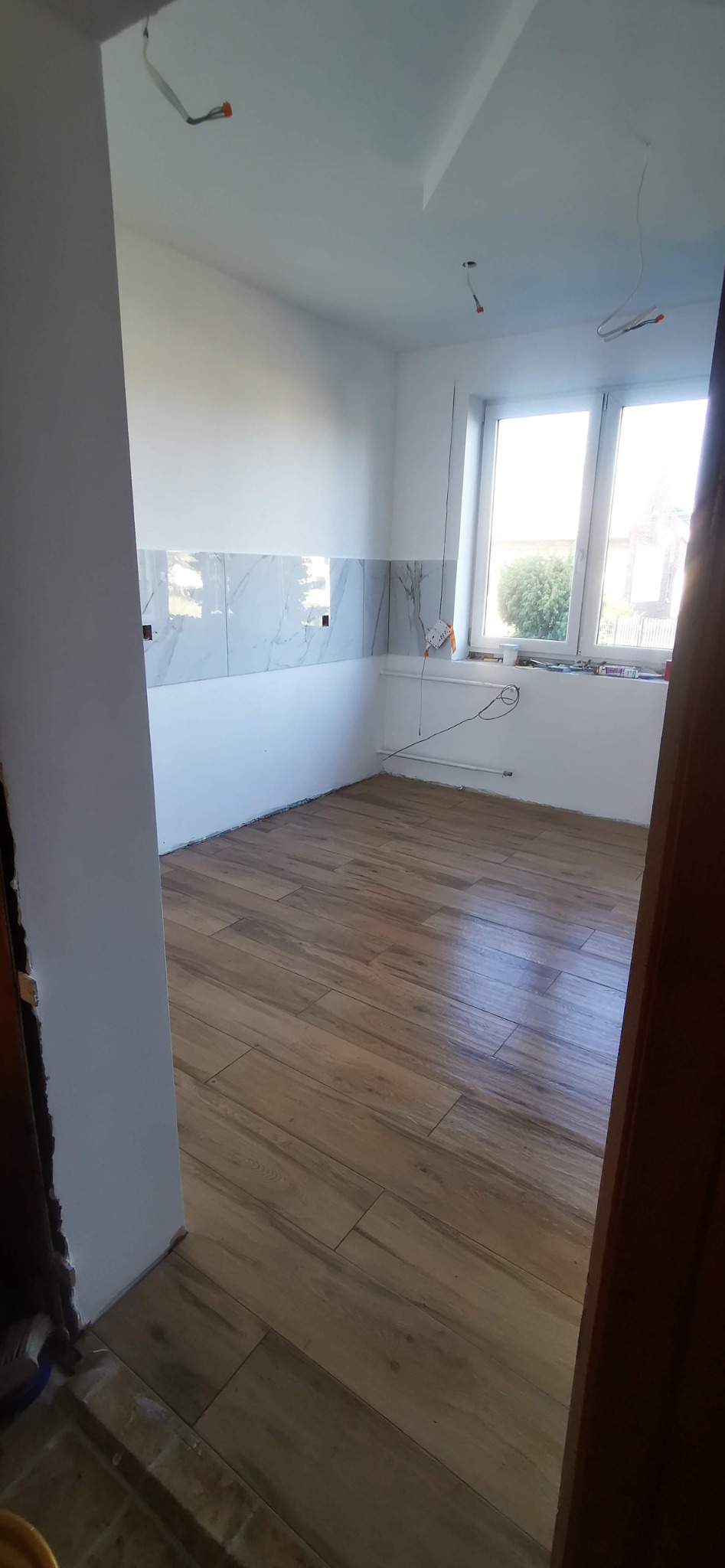Wnętrze kuchni w trakcie prac wykończeniowych, widoczne płytki imitujące marmur na ścianie, drewnopodobna podłoga, surowy stan instalacji elektrycznej.
