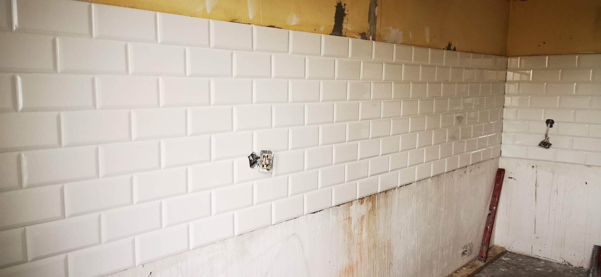 Ściana w trakcie układania białych, prostokątnych płytek imitujących cegiełki, widoczne puszki elektryczne i poziomica oparta o ścianę w rogu pomieszczenia.