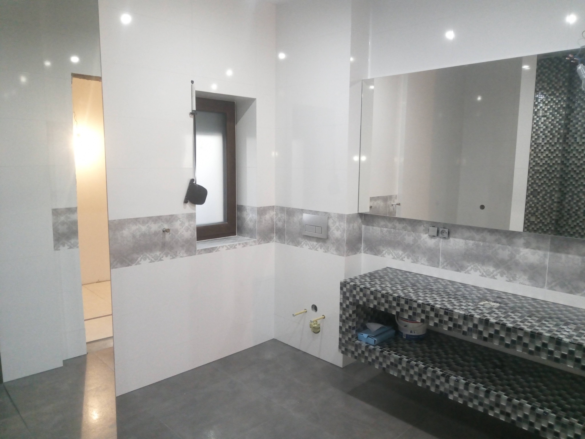 Nowoczesna łazienka z białymi płytkami, szarym pasem dekoracyjnym i umywalką z mozaiki. Widoczne okno z roletą i zamontowana podtynkowa spłuczka WC.