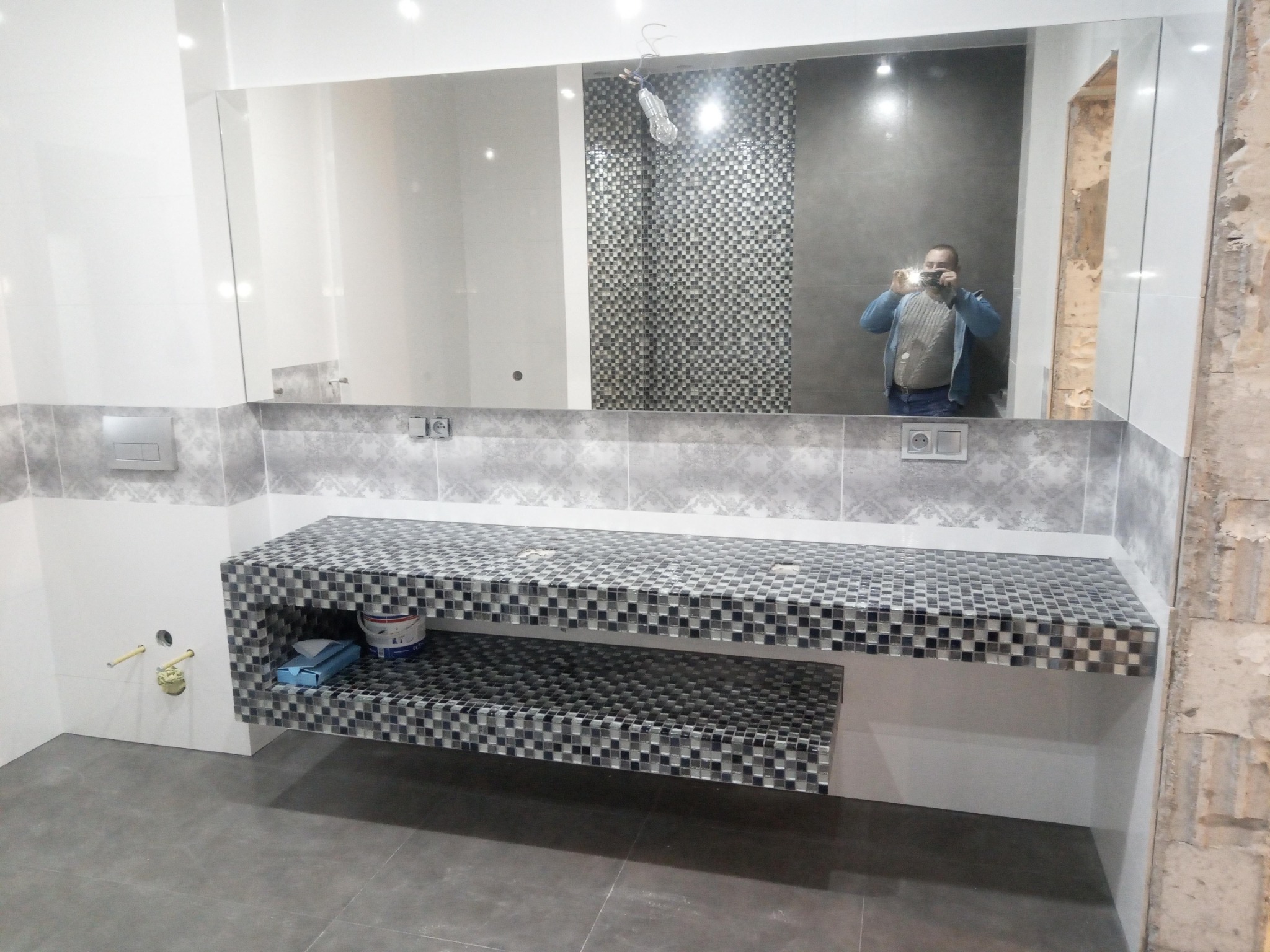 Ukończona zabudowa umywalki z mozaiki w łazience, widoczne przyłącza wodne i odbicie fotografa w lustrze.