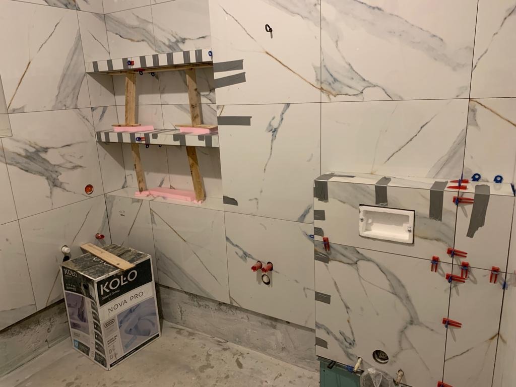 Łazienka w trakcie wykańczania: białe płytki imitujące marmur, zamontowane półki z drewnianą konstrukcją wsporczą i różowym wypełnieniem, obudowa stelaża WC w trakcie oklejania płytkami, widoczne...