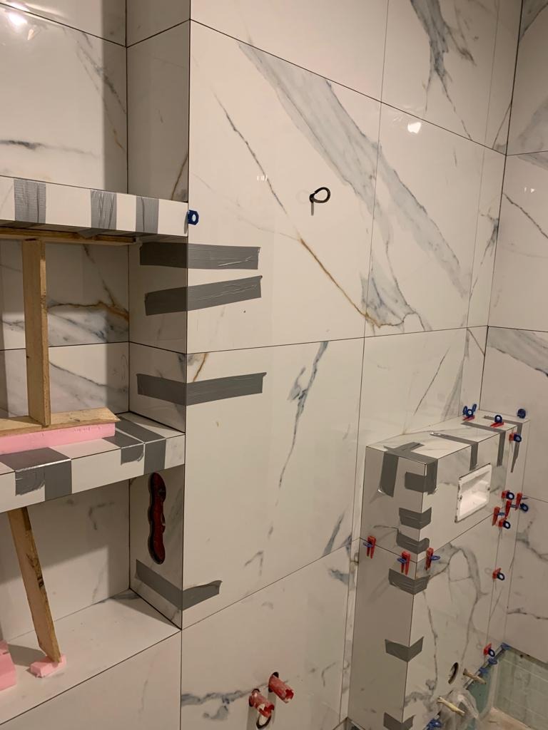 Montaż płytek imitujących marmur w łazience, widoczne elementy konstrukcyjne i mocowania, taśma maskująca zabezpieczająca krawędzie, oraz wystające rury instalacji wodnej.