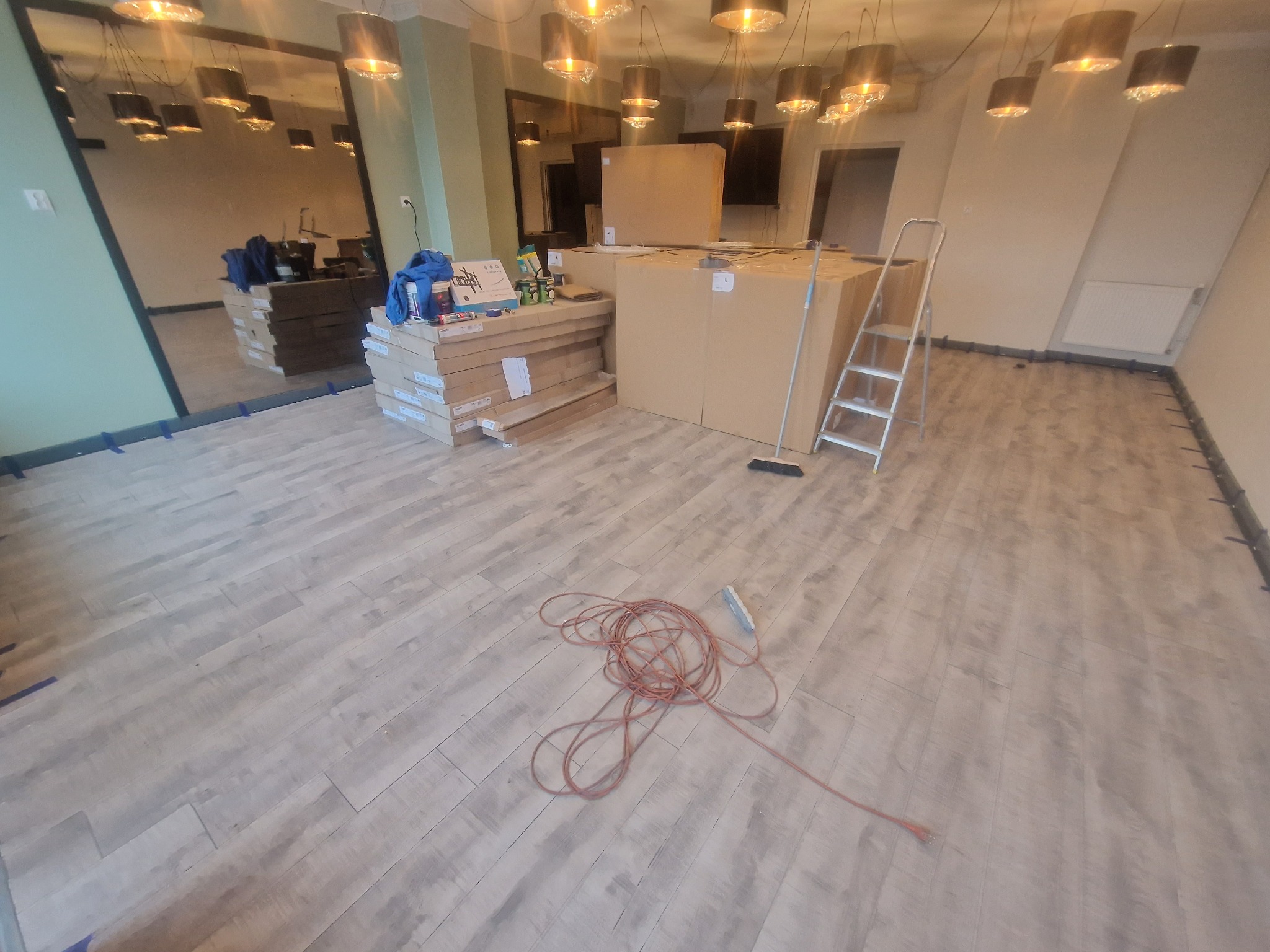 Układanie paneli podłogowych w jasnym pomieszczeniu, widoczne paczki paneli, drabina, przedłużacz oraz odbicie w lustrze