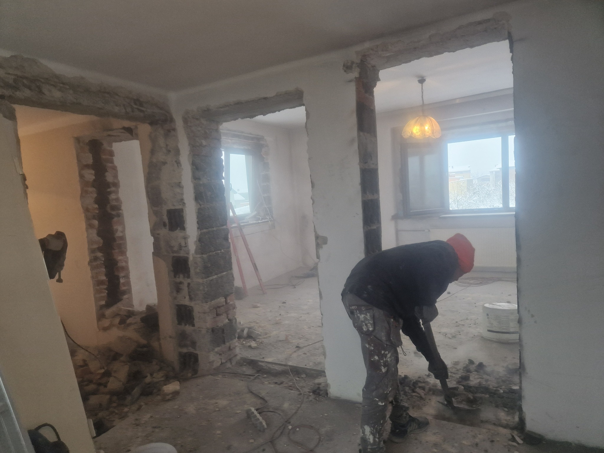Pracownik w ubrudzonych farbą spodniach, w trakcie kucia resztek ściany działowej w mieszkaniu, widoczne gruz i usunięte fragmenty muru, w tle dwa okna i drabina.