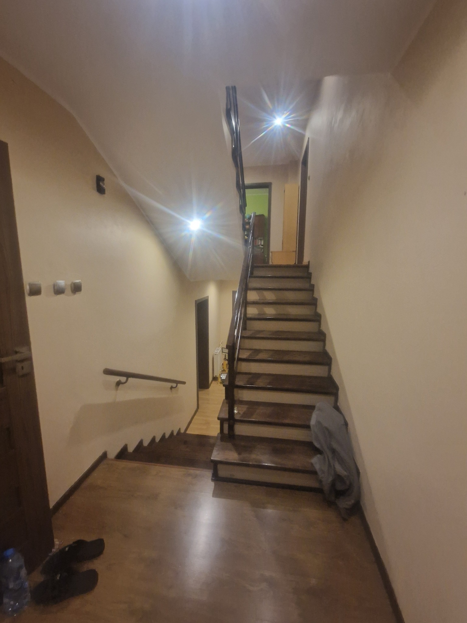 Wnętrze domu z drewnianymi schodami z metalową balustradą, widok na korytarz i drzwi, ściany w odcieniach beżu, na podłodze leżą buty i butelka wody.