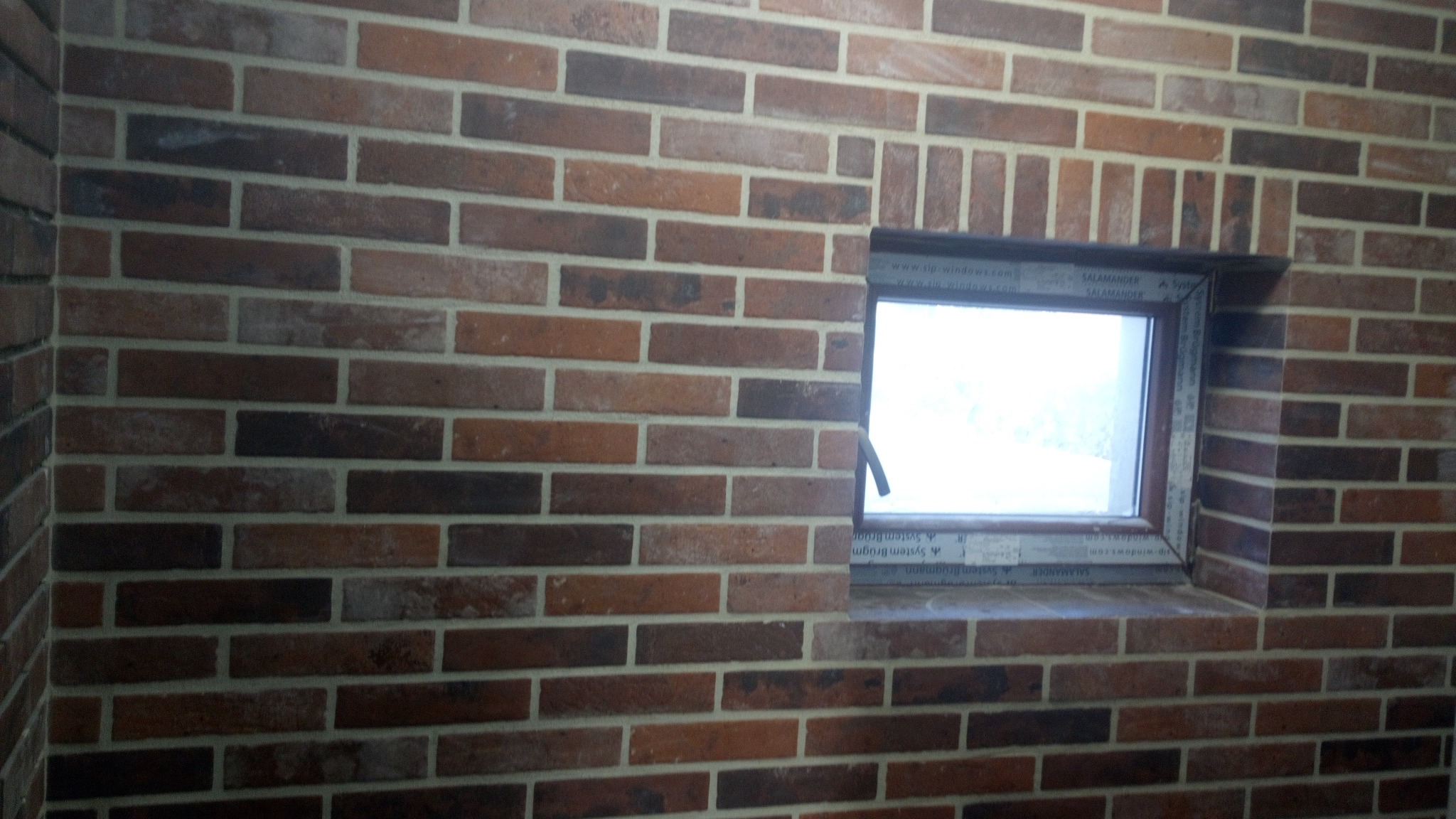 Ściana z cegły klinkierowej w odcieniach brązu i czerwieni, z wbudowanym oknem PCV z widoczną ramą. Cegły ułożone w regularny wzór z jasną fugą.