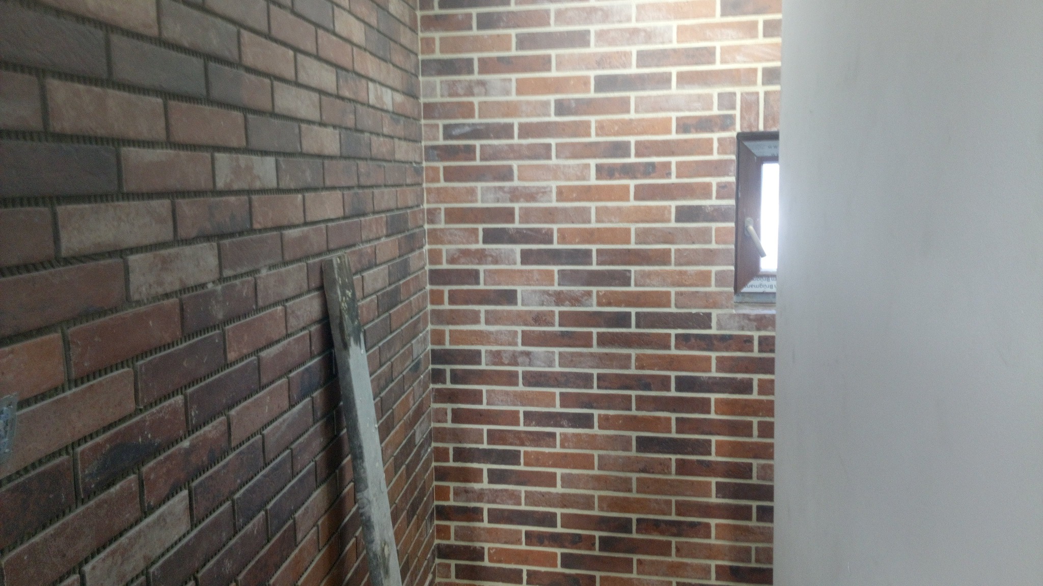 Ściana wyłożona cegiełkami w odcieniach brązu i beżu, widoczna fugowana spoiny, fragment okna i listwa przypodłogowa oparta o ścianę.