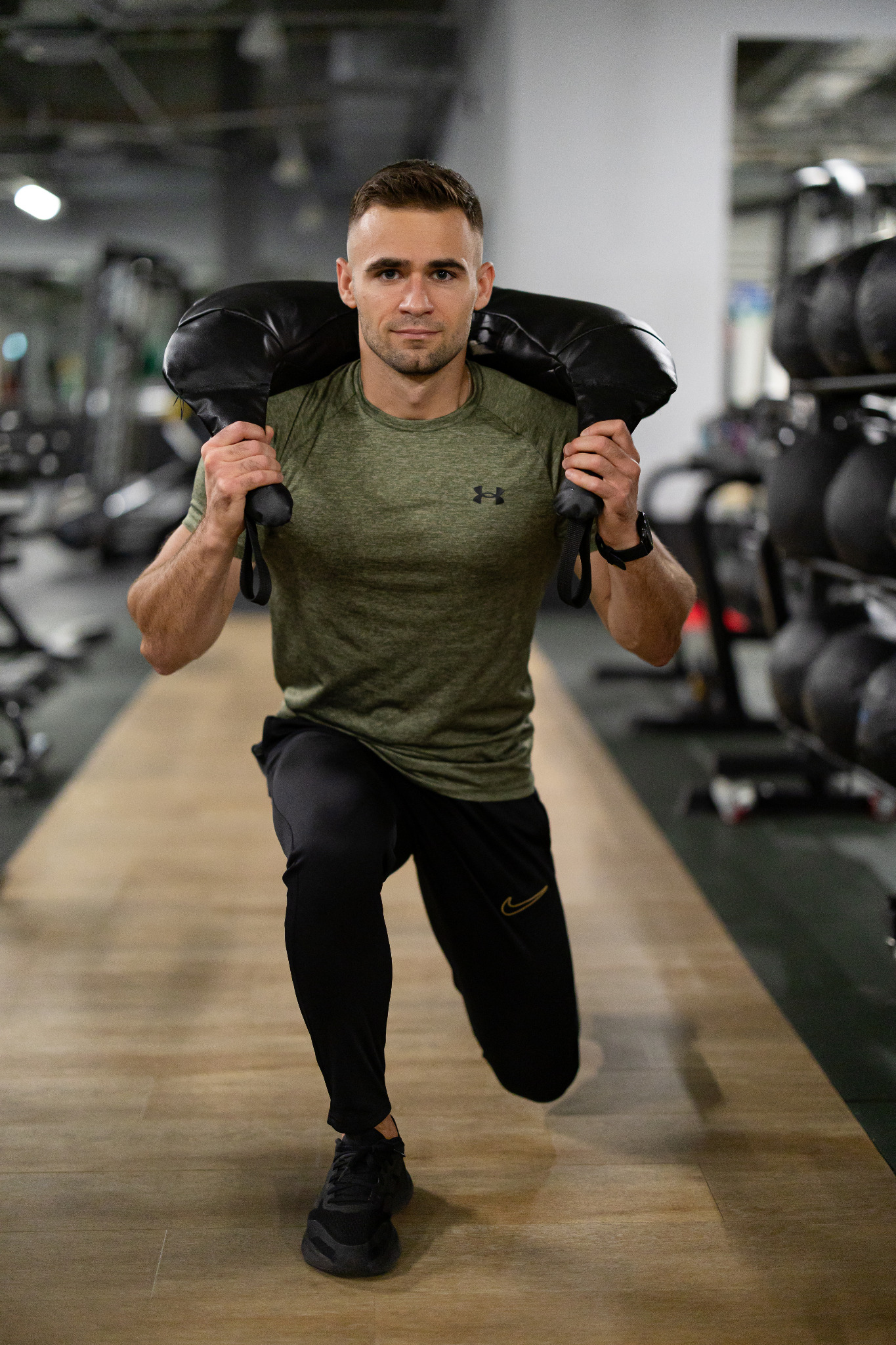 Mężczyzna w sportowym stroju wykonuje wykrok z obciążeniem na barkach w siłowni. Ćwiczenie siłowe, trening funkcjonalny, budowanie siły i kondycji.