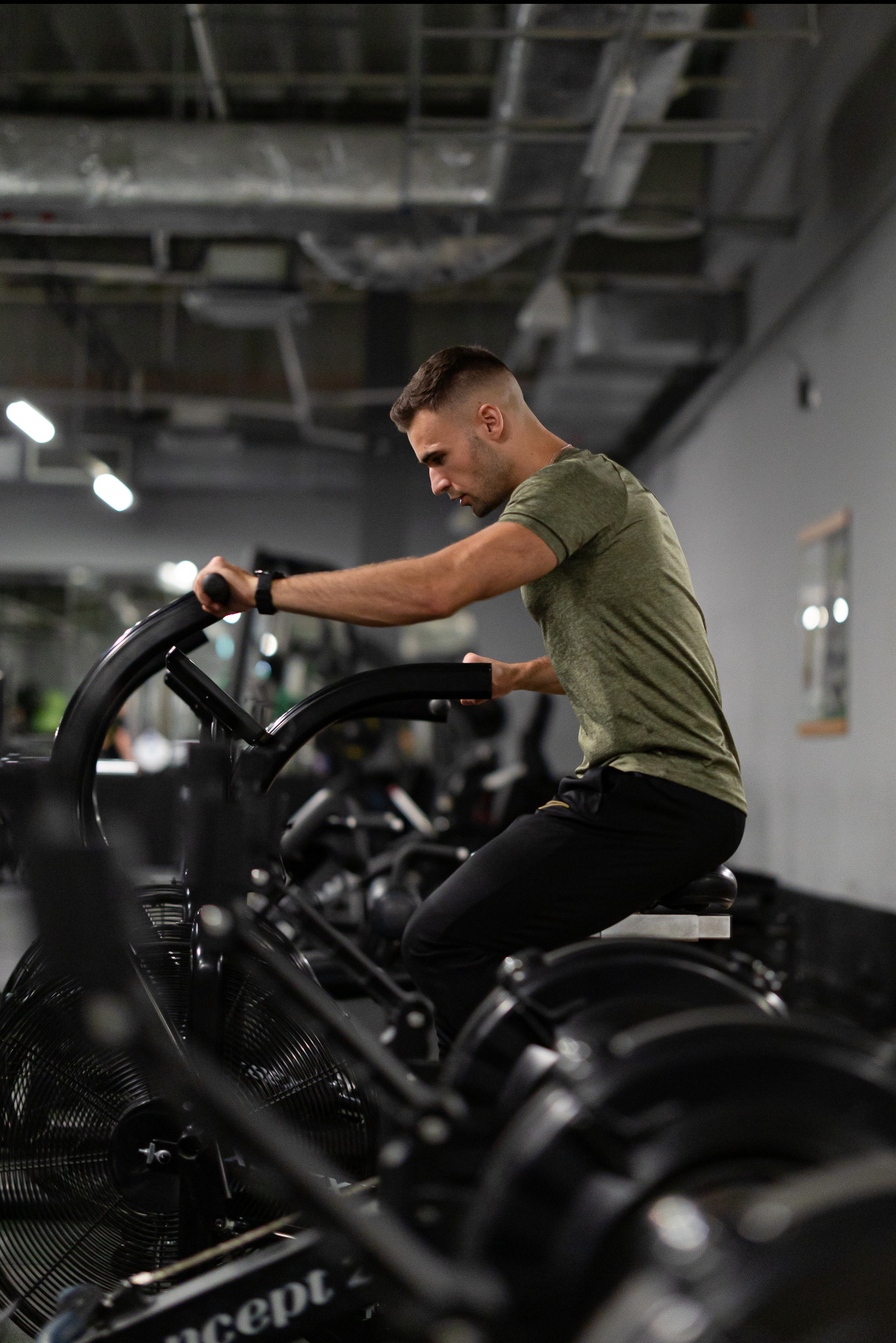 Młody mężczyzna ćwiczy na rowerze powietrznym w siłowni, ubrany w zieloną koszulkę i czarne spodnie. Widoczne inne maszyny i oświetlenie sufitowe. Intensywny trening.