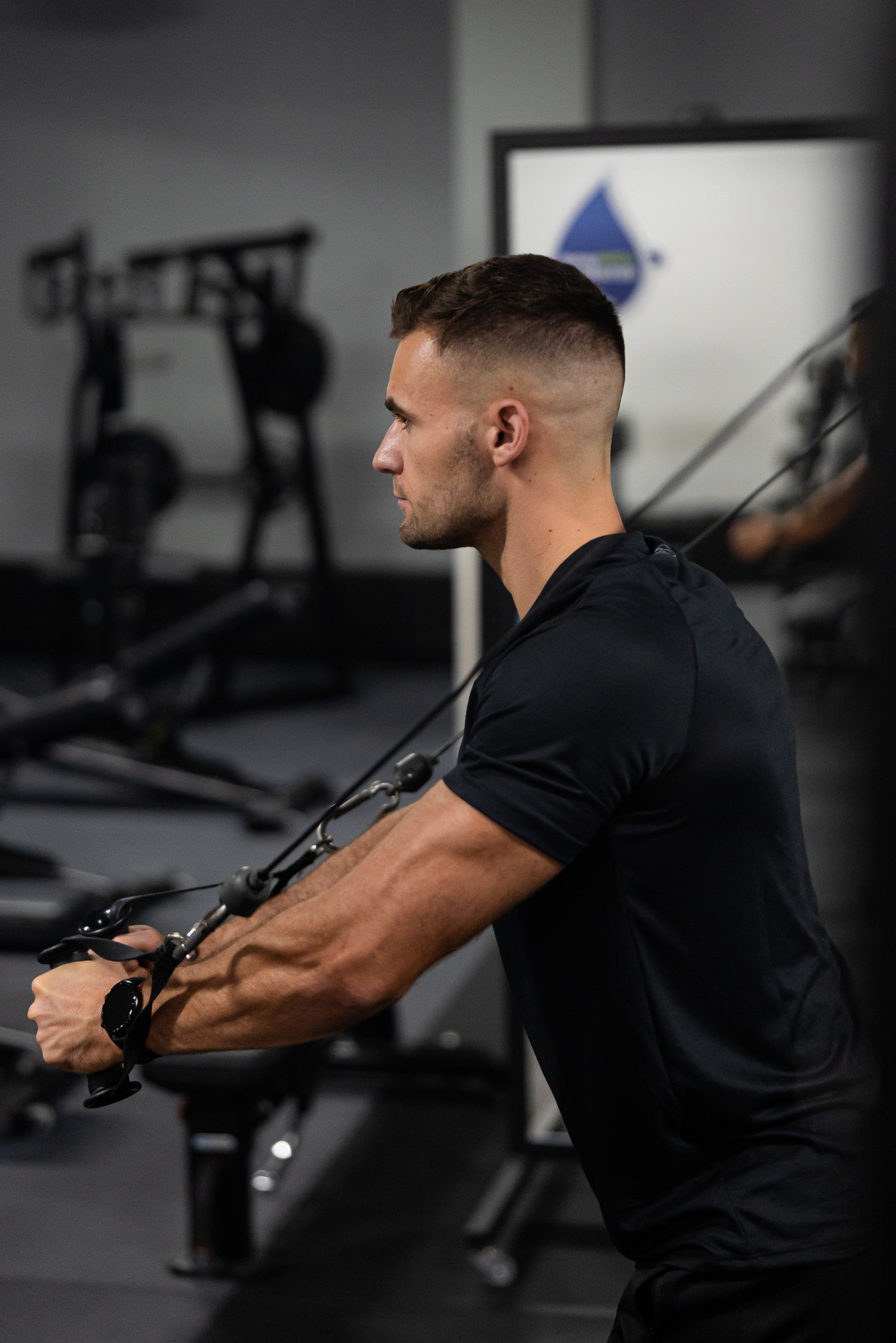 Mężczyzna w czarnej koszulce ćwiczy na maszynie w siłowni, profil, widoczne napięte mięśnie ramion, w tle sprzęt do ćwiczeń i logo na ścianie.