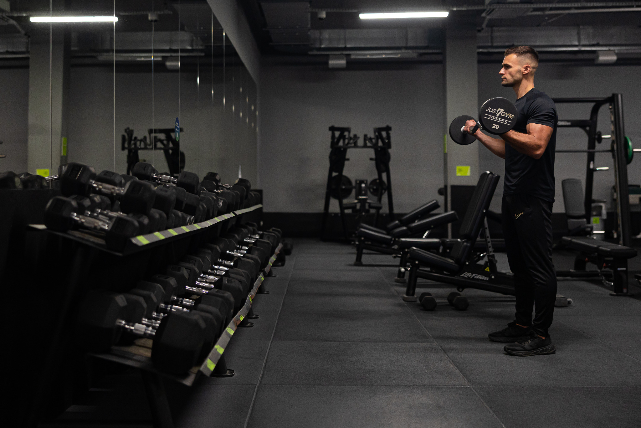 Mężczyzna w czarnym stroju ćwiczy biceps z obciążeniem 20 kg w siłowni Just Gym, obok regał z hantlami i sprzęt do ćwiczeń. Profil, oświetlenie górne.