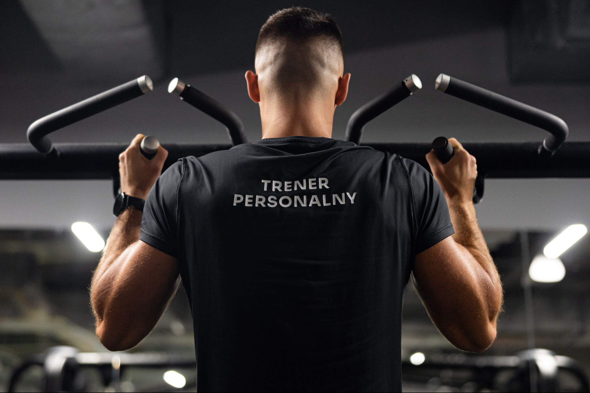 Mężczyzna ćwiczy na drążku do podciągania, na koszulce napis 'TRENER PERSONALNY'. Widok z tyłu, skupienie na muskulaturze ramion i pleców.