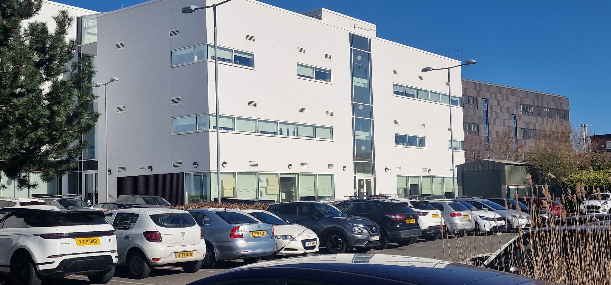 Biały budynek biurowy z rzędami okien, oświetlony słońcem, z zaparkowanymi samochodami. Widok na elewację i otoczenie budynku w słoneczny dzień.