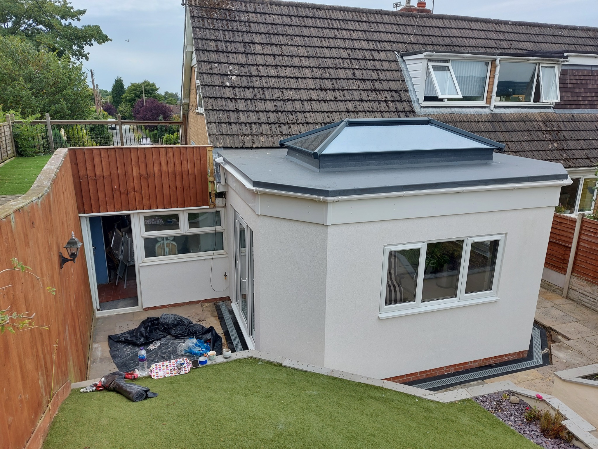 Wykończona, jasna elewacja dobudówki z płaskim dachem i świetlikiem. Widoczne okna PCV, fragment ogrodu i domu z brązową dachówką.