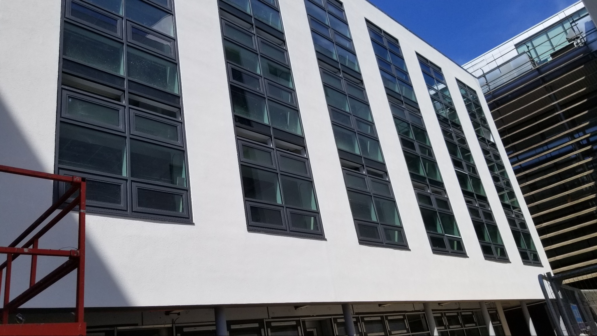 Biała elewacja budynku z rzędami okien w ciemnych ramach. Fragment rusztowania po lewej stronie. Widok z dołu, błękitne niebo w tle.