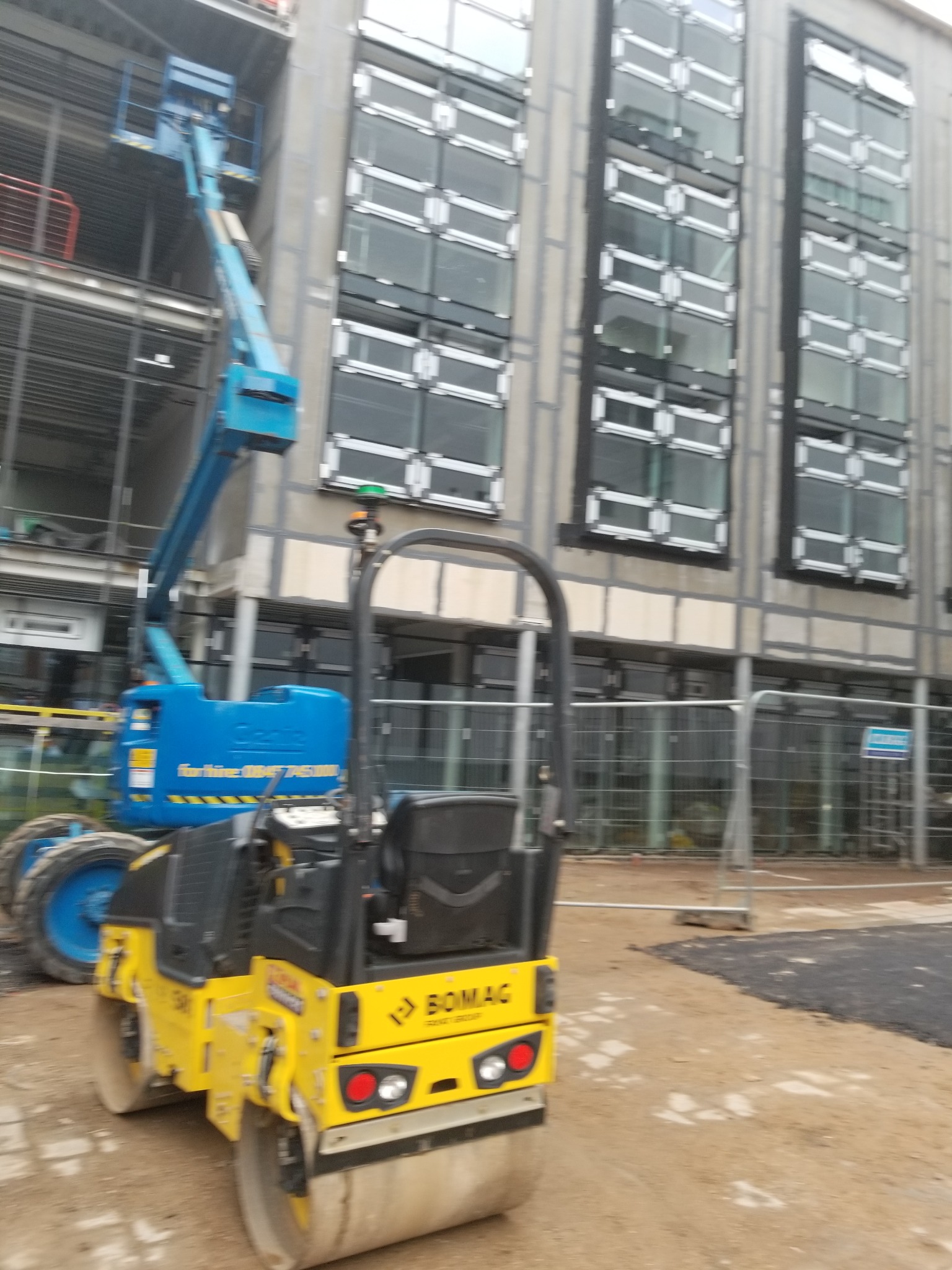 Żółty walec Bomag na placu budowy, w tle budynek z oknami i podnośnik Genie. Prace ziemne w toku, teren zabezpieczony barierkami.