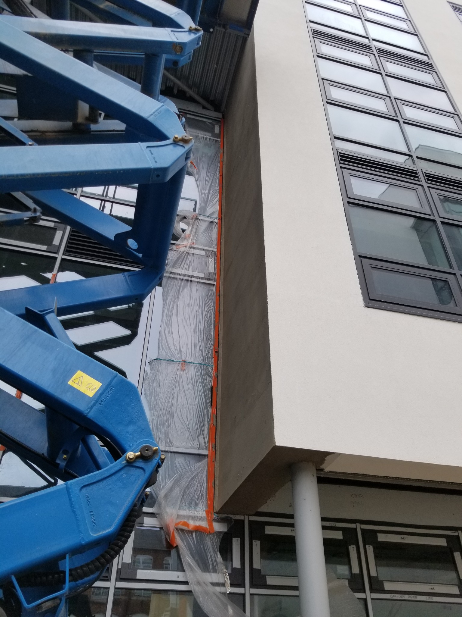 Prace elewacyjne na wysokościach z użyciem podnośnika koszowego. Fragment ocieplonej ściany budynku z oknami, zabezpieczony folią. Elementy konstrukcji podnośnika w kadrze.