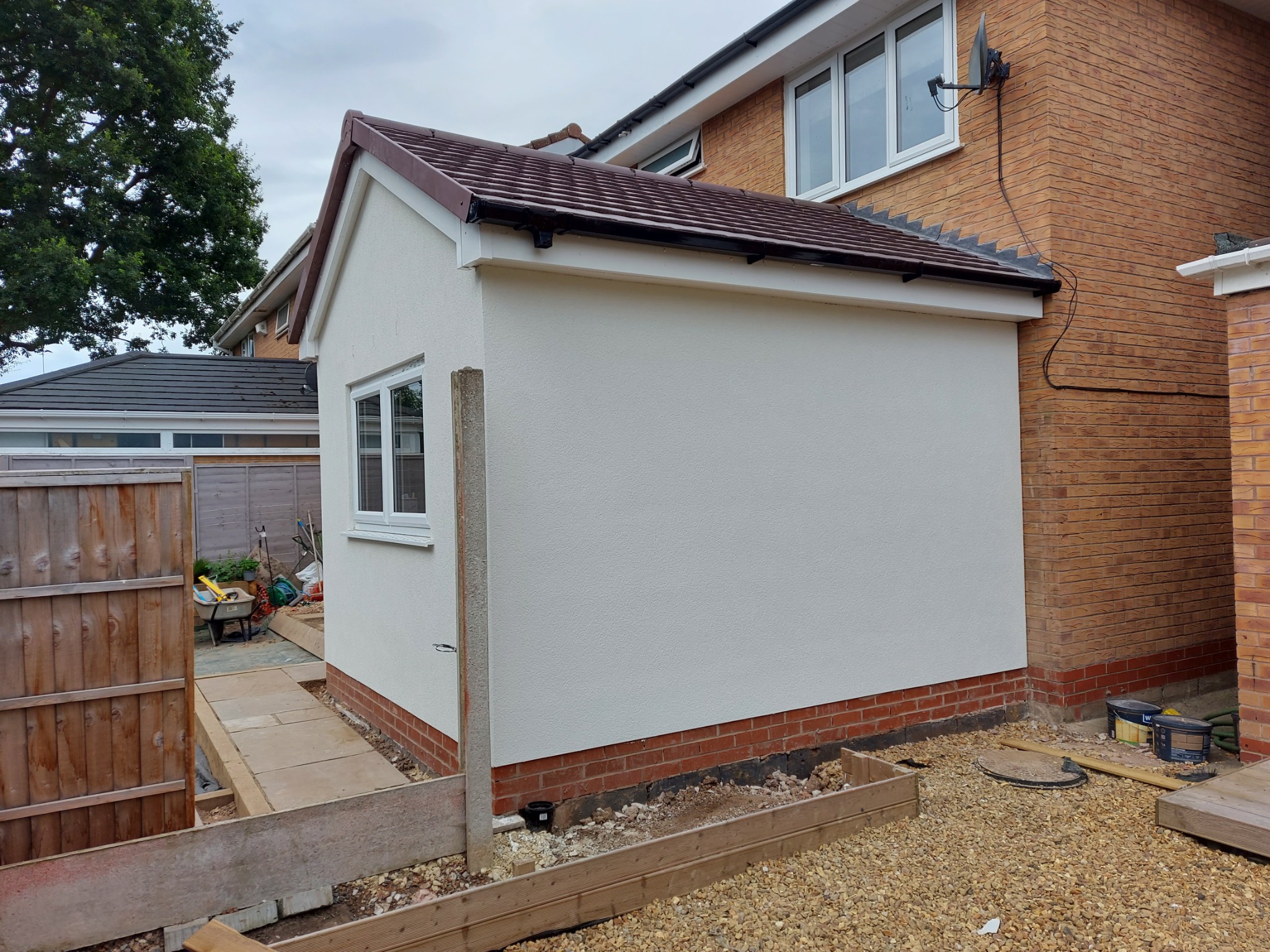 Biała, otynkowana dobudówka z brązowym dachem, przylegająca do ceglanego domu. Widoczny fragment ogrodu z elementami przygotowania pod dalsze prace wykończeniowe.