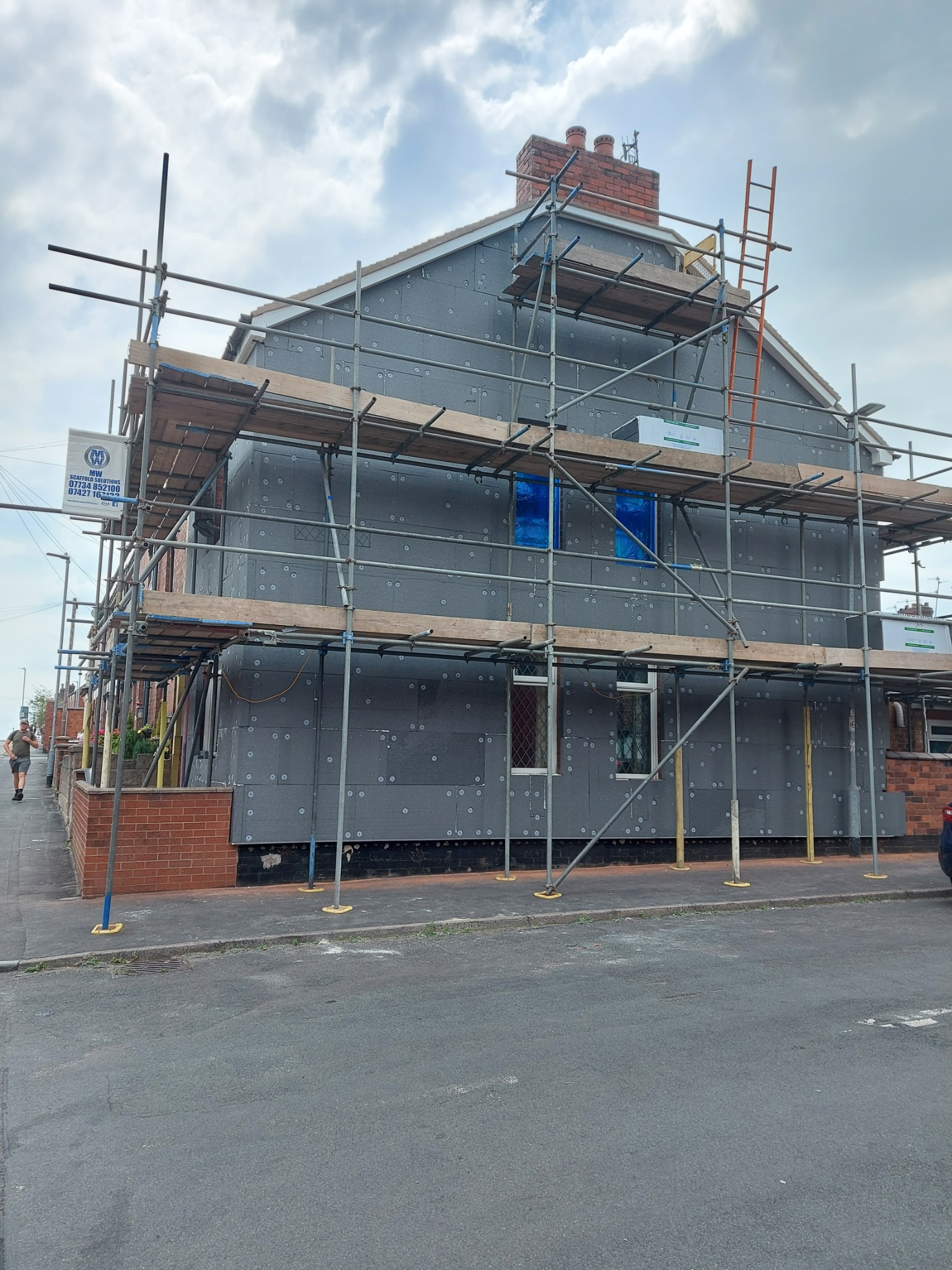 Dom jednorodzinny w trakcie ocieplania, zabezpieczony rusztowaniem. Widoczne szare płyty izolacyjne na elewacji, okna zabezpieczone folią, ceglana podmurówka.