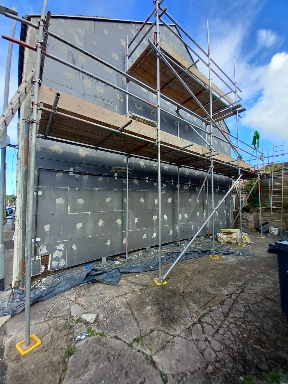 Dom w trakcie ocieplania: rusztowanie, szare płyty izolacyjne z białymi punktami mocowania, niebieskie niebo. Etap prac na zewnątrz budynku.