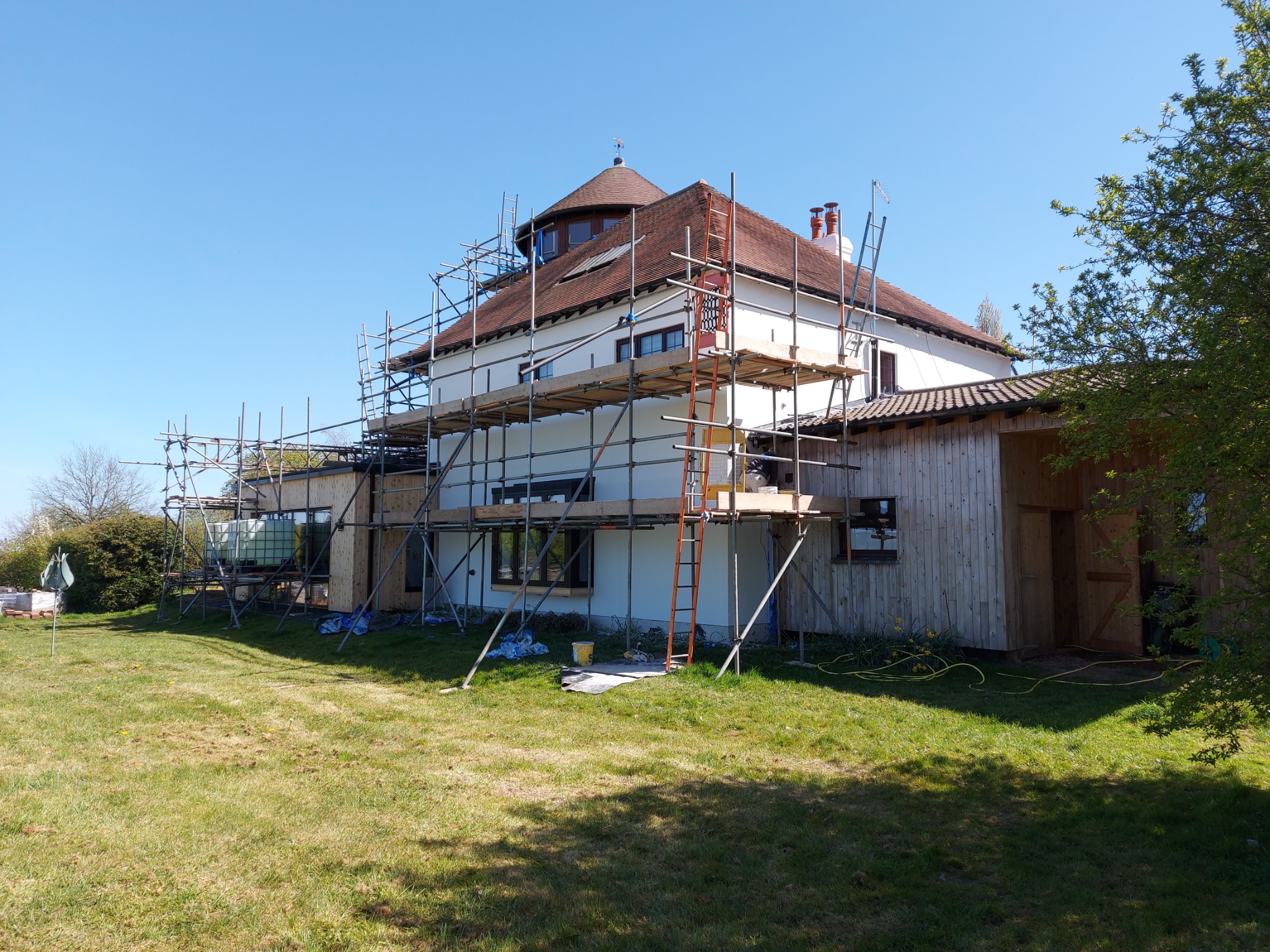 Dom jednorodzinny w trakcie prac elewacyjnych, otoczony rusztowaniami. Widoczna drabina oparta o ścianę, trawnik i fragment budynku gospodarczego z drewnianą elewacją.
