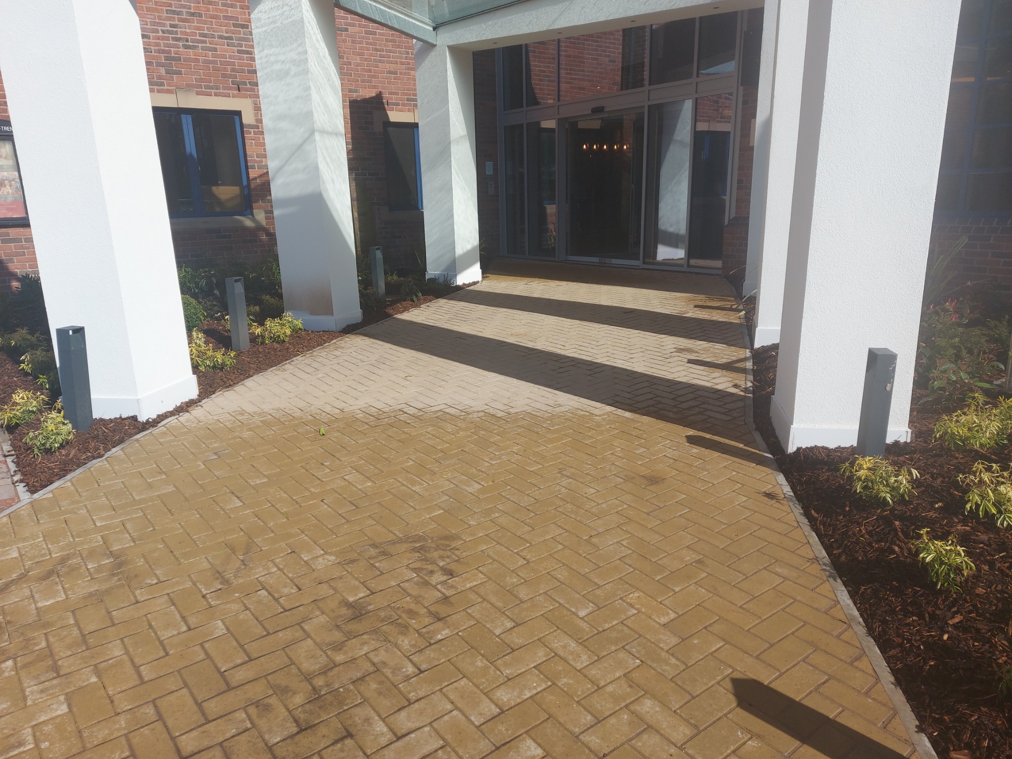 Wejście do budynku z automatycznymi drzwiami, wyłożone kostką brukową w odcieniach beżu, z białymi kolumnami i rabatami z roślinami po bokach.