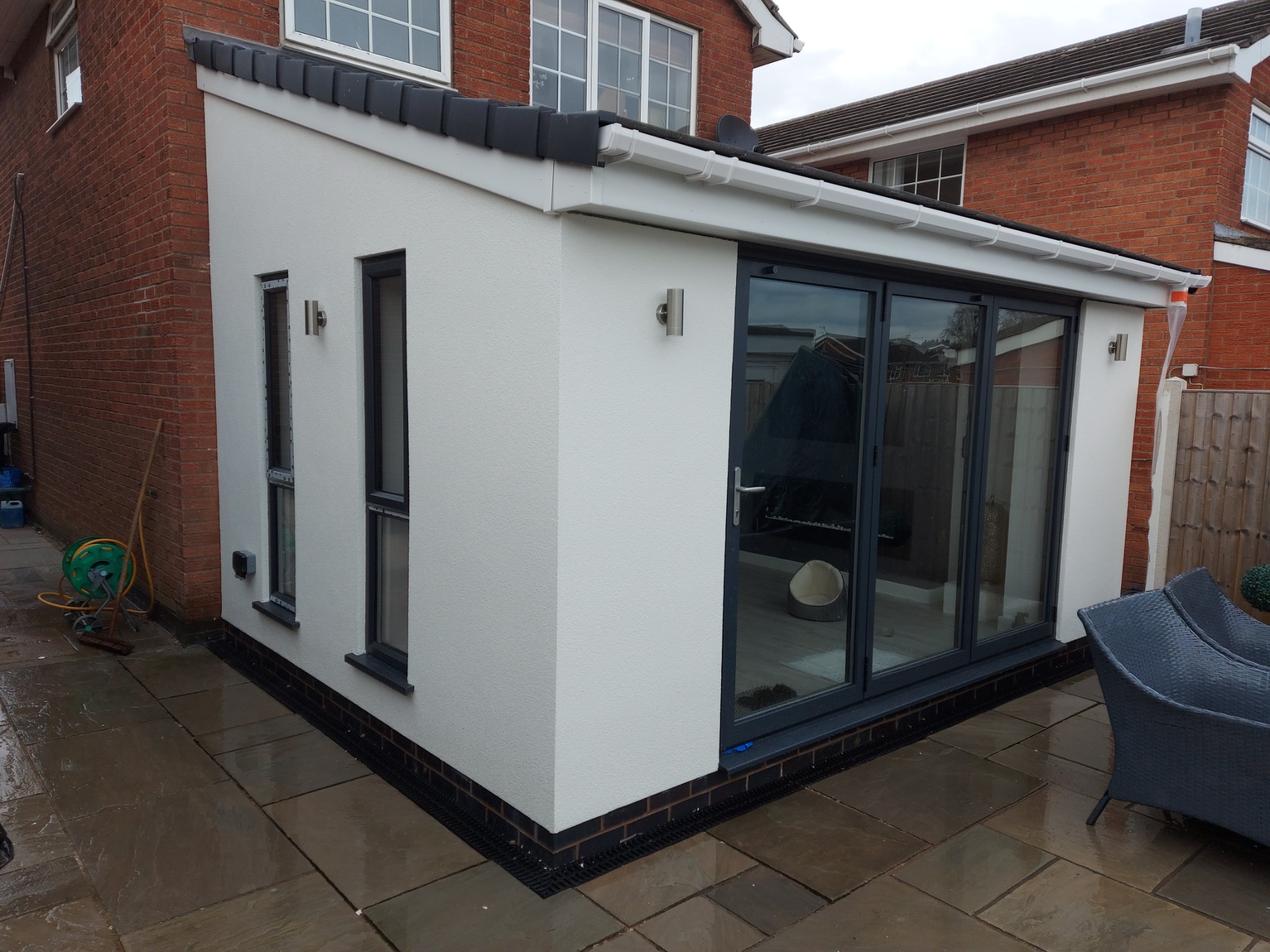 Dobudówka domu z białymi ścianami o fakturze baranka i grafitowymi oknami, widoczna z zewnątrz. Na tarasie brązowe płytki i grafitowe meble ogrodowe.