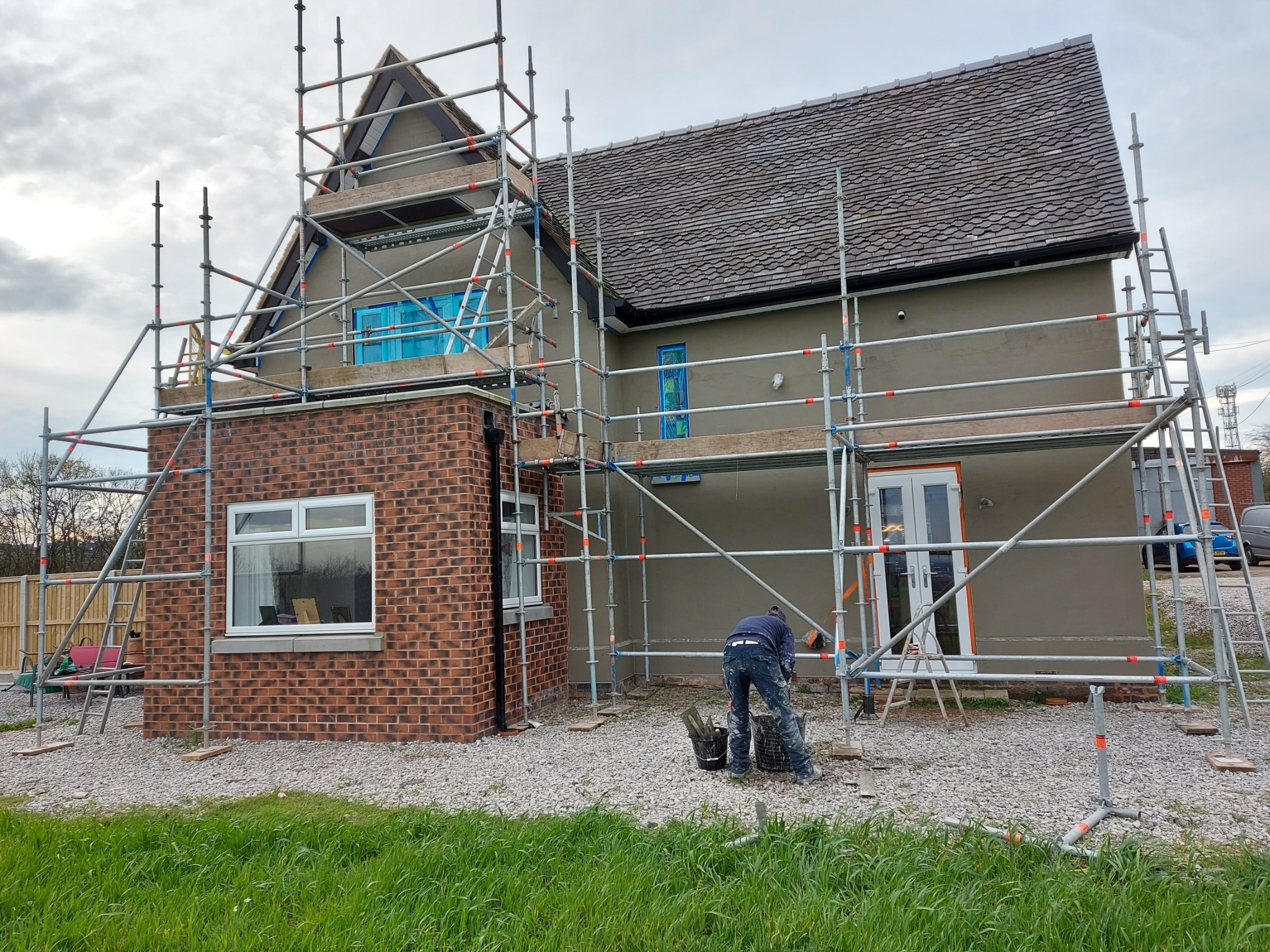 Dom w trakcie tynkowania elewacji, otoczony rusztowaniem. Widoczny pracownik przygotowujący zaprawę. Okna zabezpieczone folią, ściany pokryte szarym tynkiem.