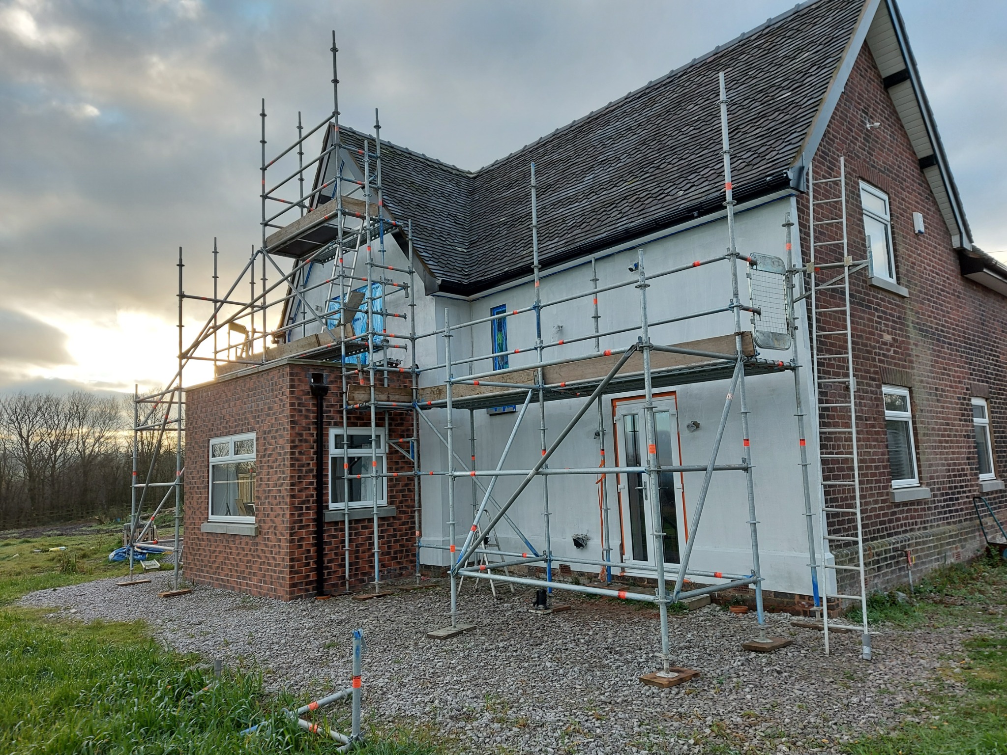 Dom z cegły i otynkowaną, białą elewacją w trakcie prac. Widoczne rusztowania, okna i fragment dachu.  Prace elewacyjne w toku, widok na dom z zewnątrz.