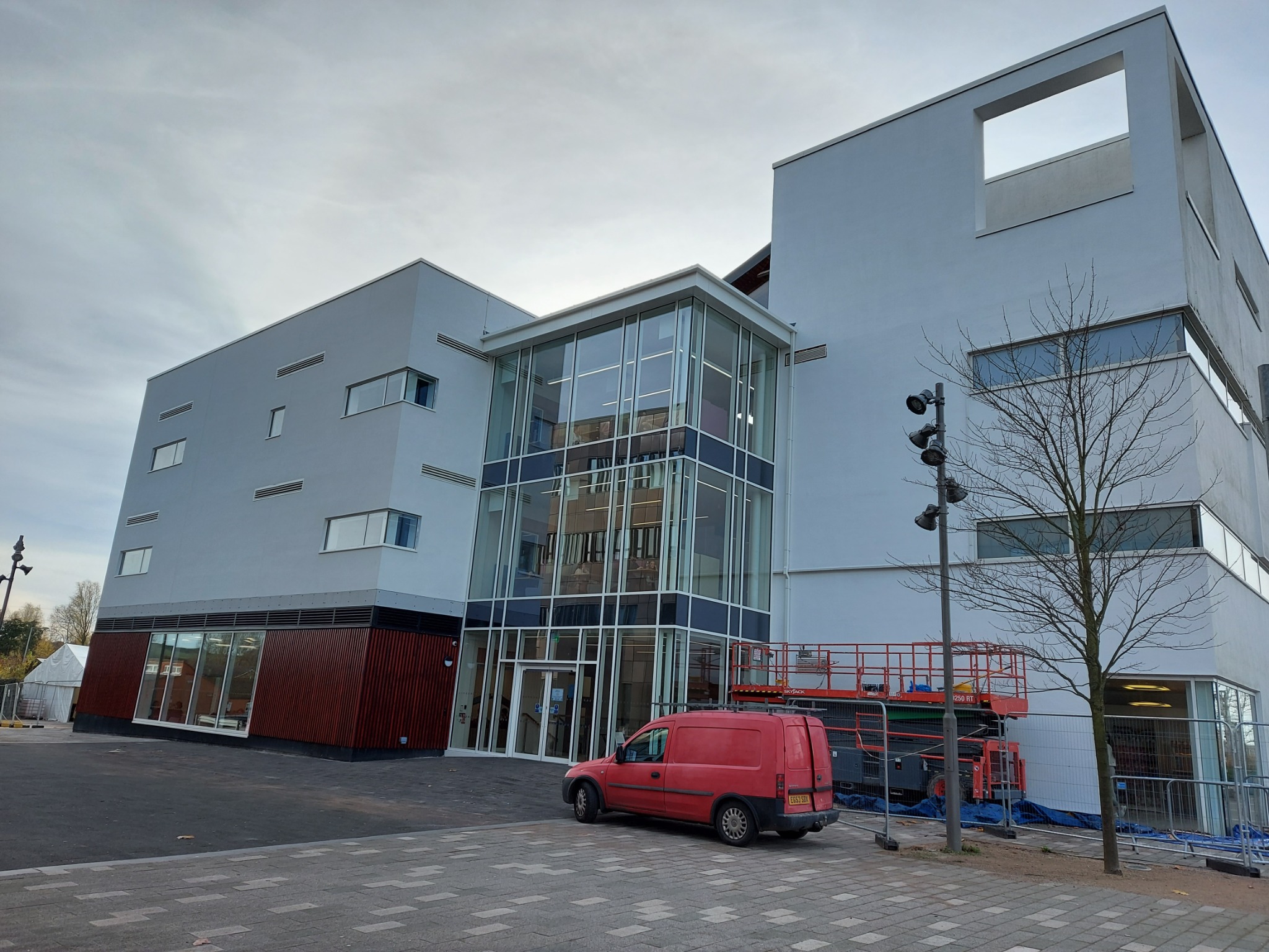 Nowoczesny budynek z przeszklonym wejściem, czerwony van zaparkowany przed nim, platforma robocza z tyłu. Minimalistyczna architektura, szara elewacja, elementy drewniane.