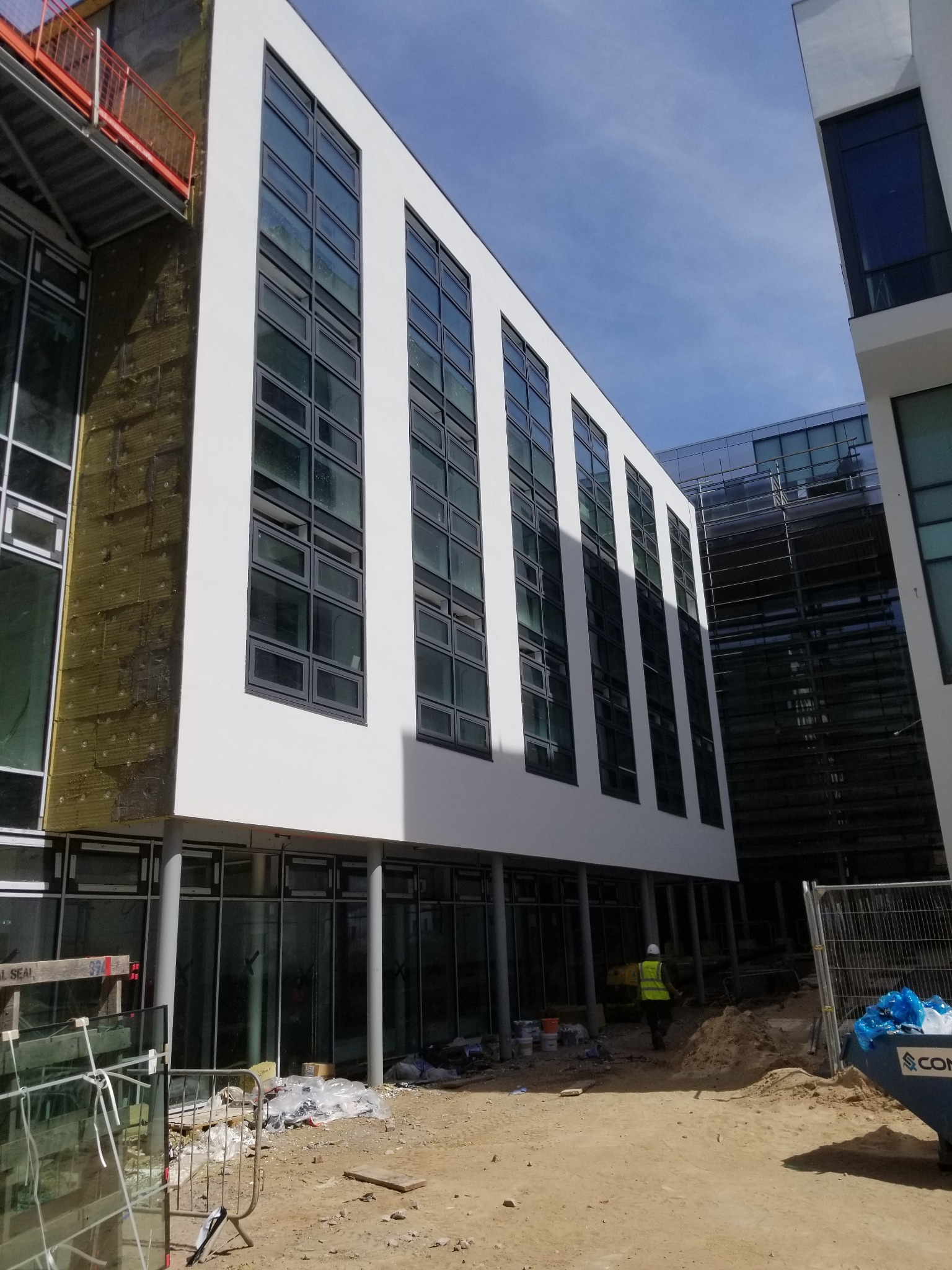 Elewacja budynku w trakcie ocieplania, widoczne fragmenty izolacji termicznej. Pracownik w odblaskowej kamizelce na terenie budowy. Budynek z licznymi oknami.