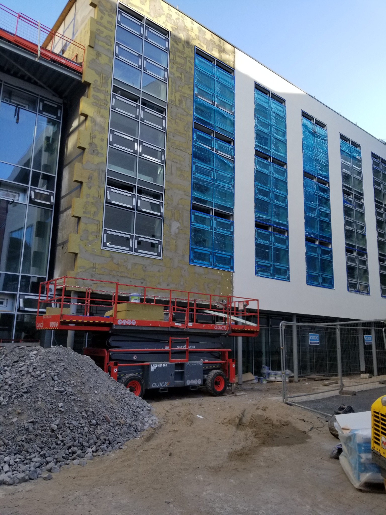 Ocieplanie budynku wielorodzinnego z użyciem podnośnika. Część elewacji ocieplona, okna zabezpieczone folią. Na dole hałda gruzu i piasku.