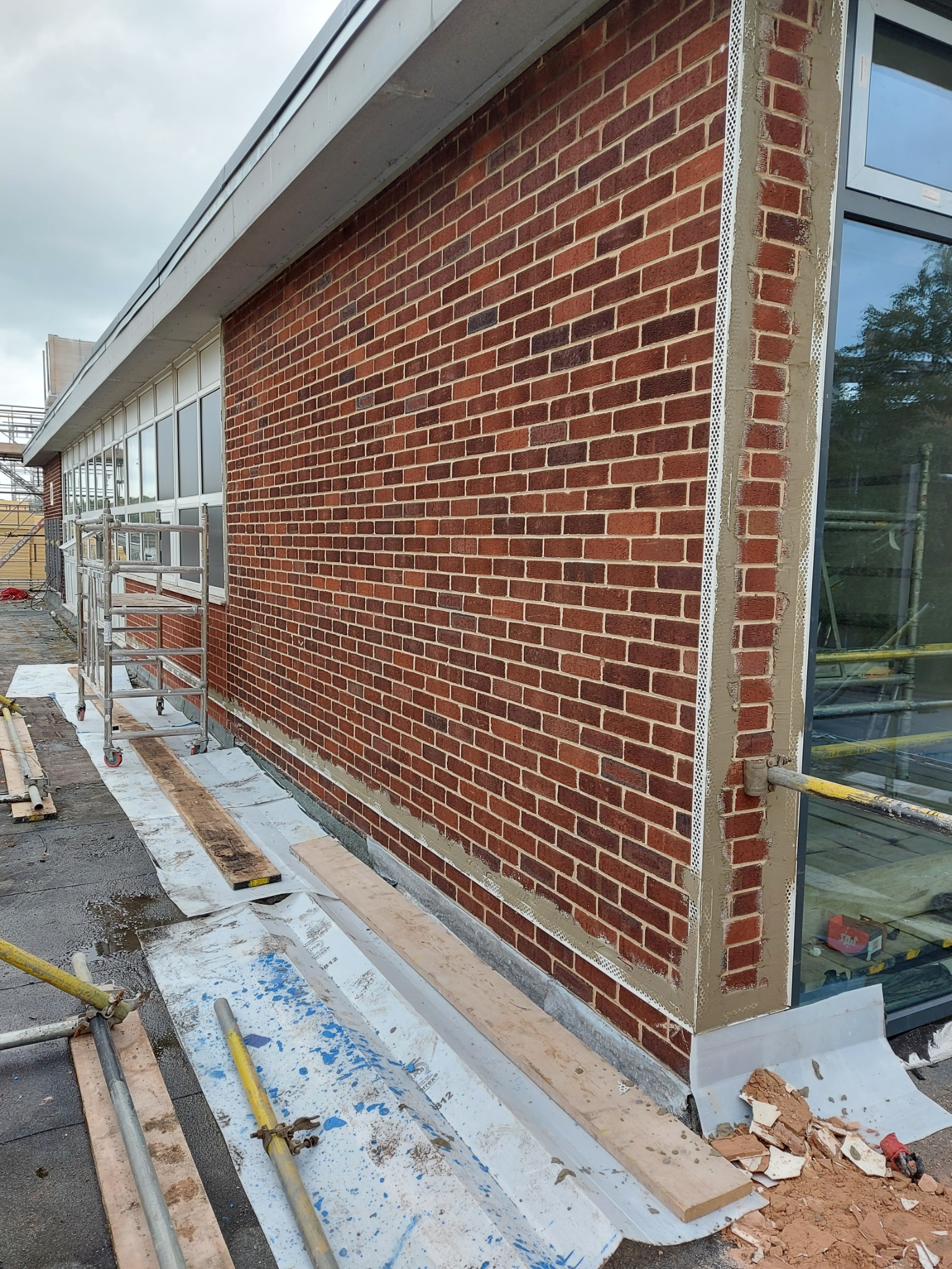 Ceglana ściana budynku w trakcie prac elewacyjnych. Widoczne profile narożnikowe i fragment rusztowania. Prace przygotowawcze do tynkowania.