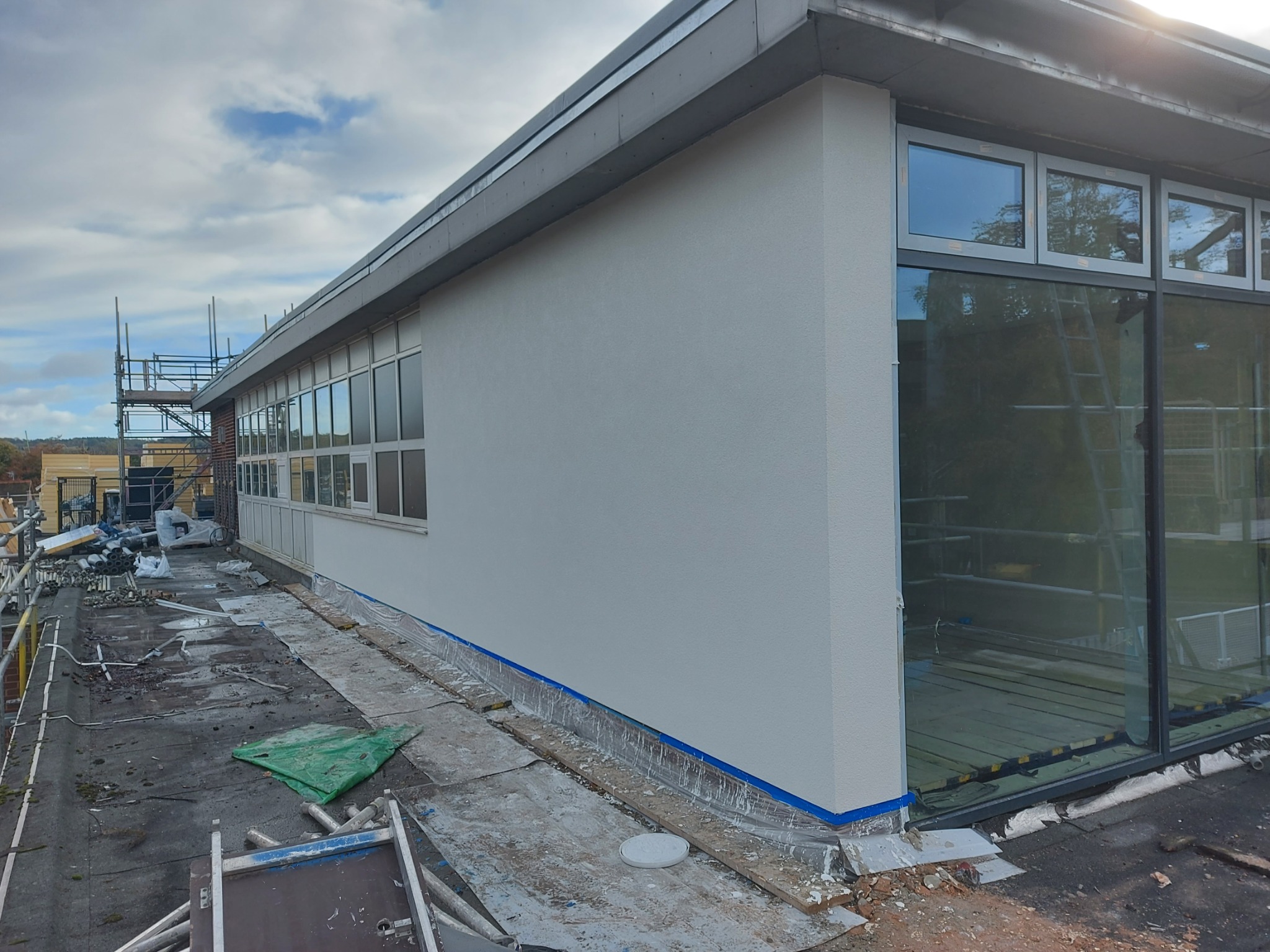 Świeżo otynkowana, biała ściana budynku z rzędem okien, zabezpieczona niebieską taśmą malarską. Na pierwszym planie widoczne materiały budowlane i rusztowanie.