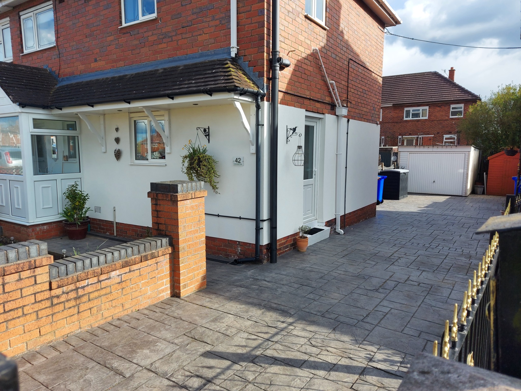 Odnowiona elewacja domu z czerwonej cegły z białymi ścianami, dekoracjami i zadbanym otoczeniem. Widoczny fragment kostki brukowej i ogrodzenie.