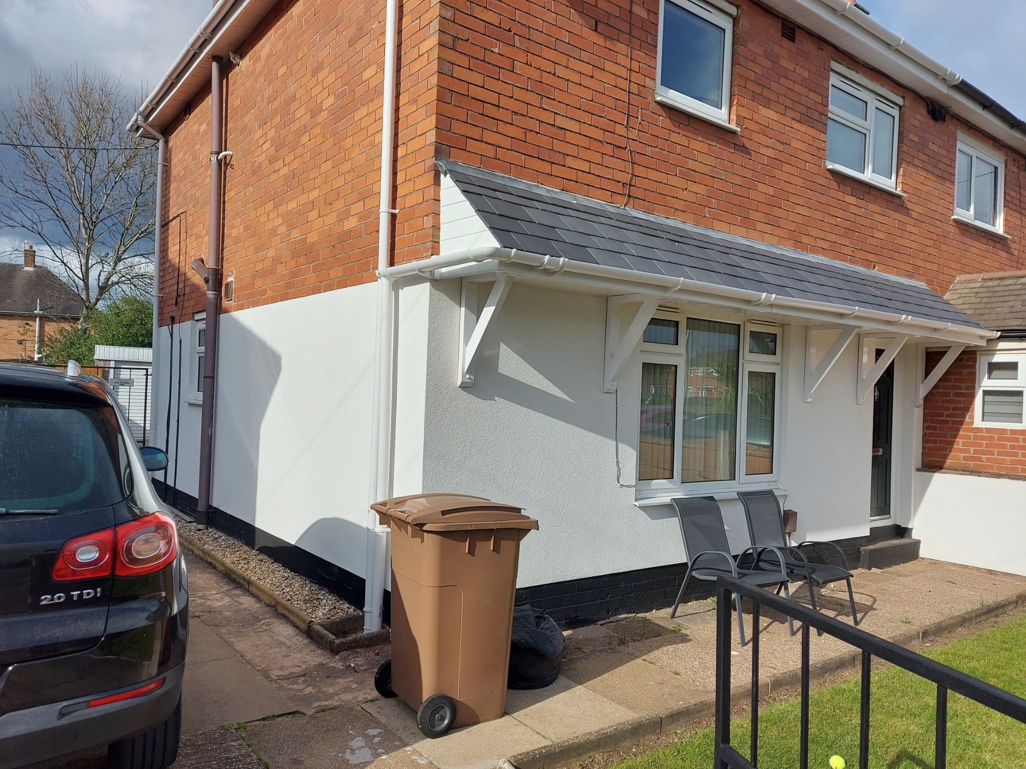 Odnowiona elewacja domu z cegły, z pomalowaną na biało dolną częścią i widoczną rynną. Na pierwszym planie brązowy kosz na śmieci i fragment czarnego samochodu.