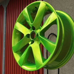 POLEROWANIE FELG Marcin Górski - Felga samochodowa po renowacji, pokryta jaskrawym, zielonym lakierem z efektem połysku, zawieszona na stojaku na tle ściany z blachy trapezowej.