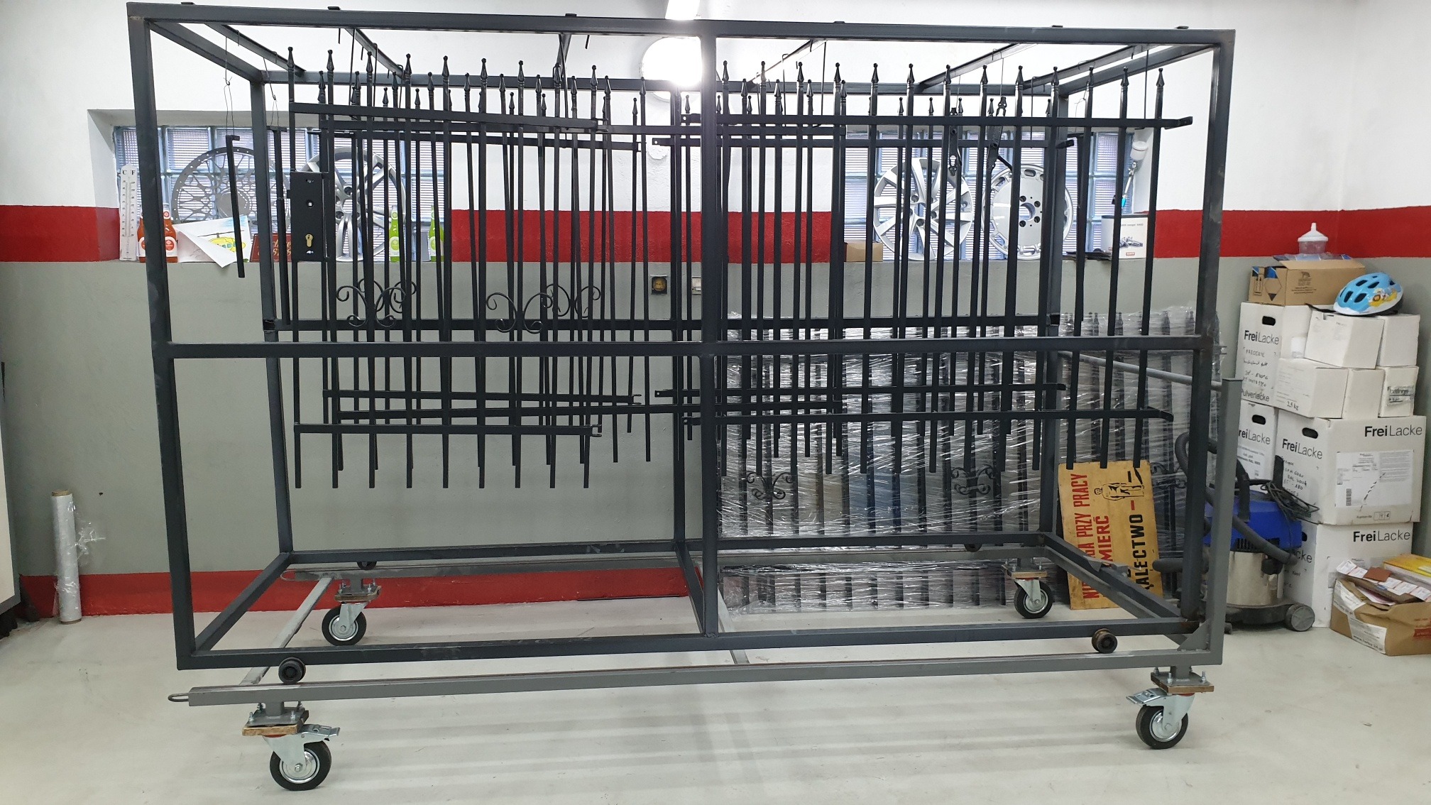 Metalowa konstrukcja na kółkach, wypełniona elementami balustrady, prawdopodobnie po procesie malowania, widoczne opakowania farb proszkowych w tle.