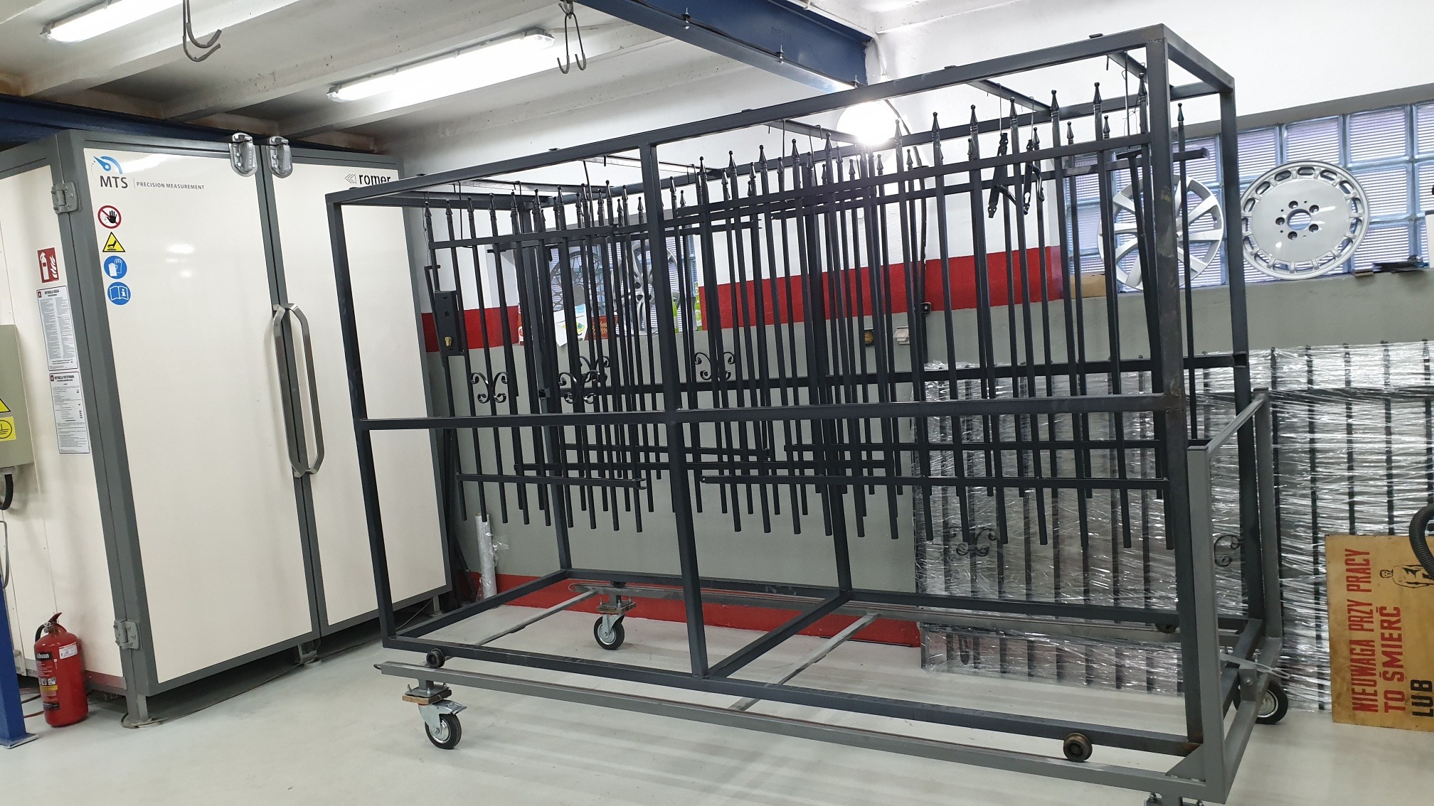 Metalowy wózek na kółkach z zawieszonymi elementami metalowymi przed piecem do malowania proszkowego w warsztacie.