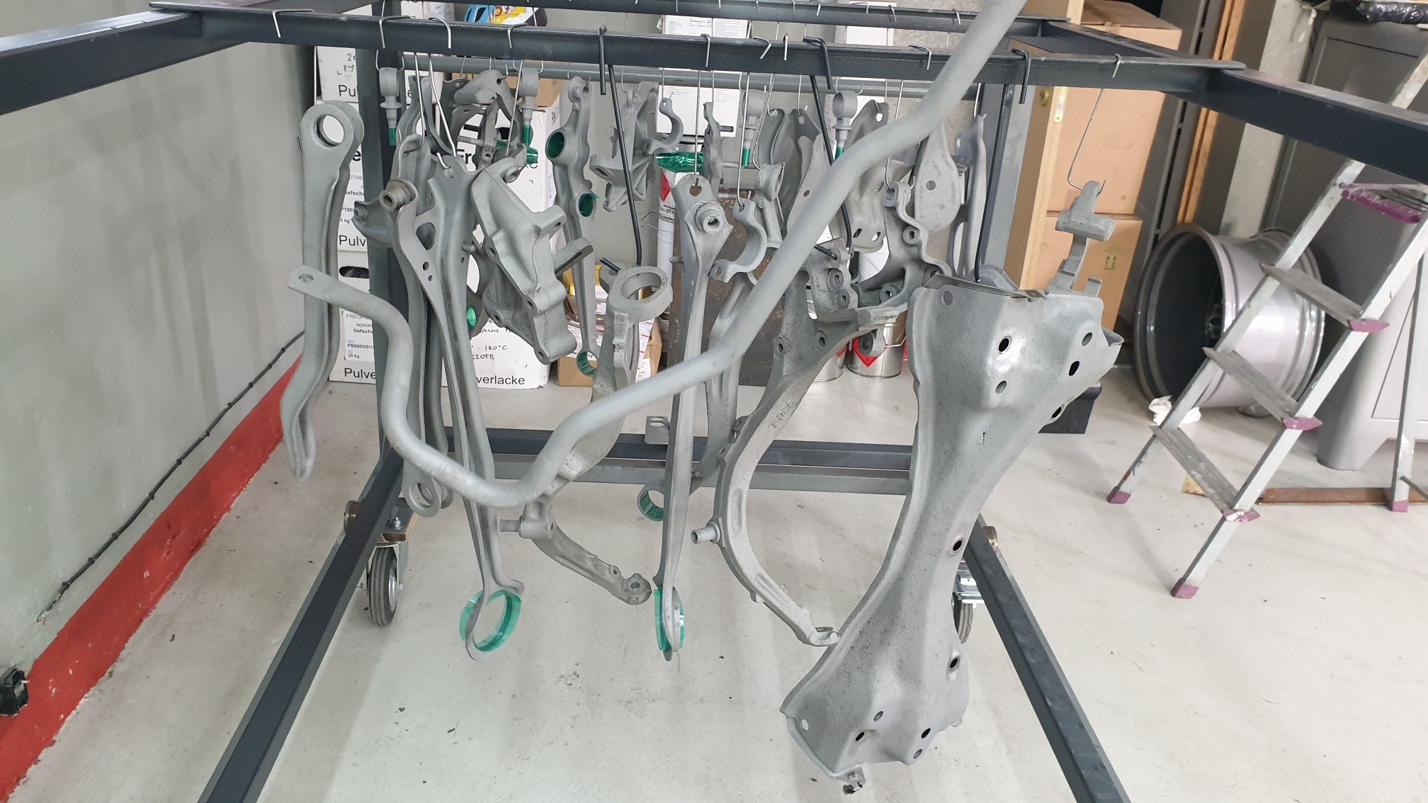 Metalowe elementy zawieszenia samochodowego po obróbce, zawieszone na wieszakach w hali produkcyjnej, przygotowane do dalszej obróbki lub wysyłki.