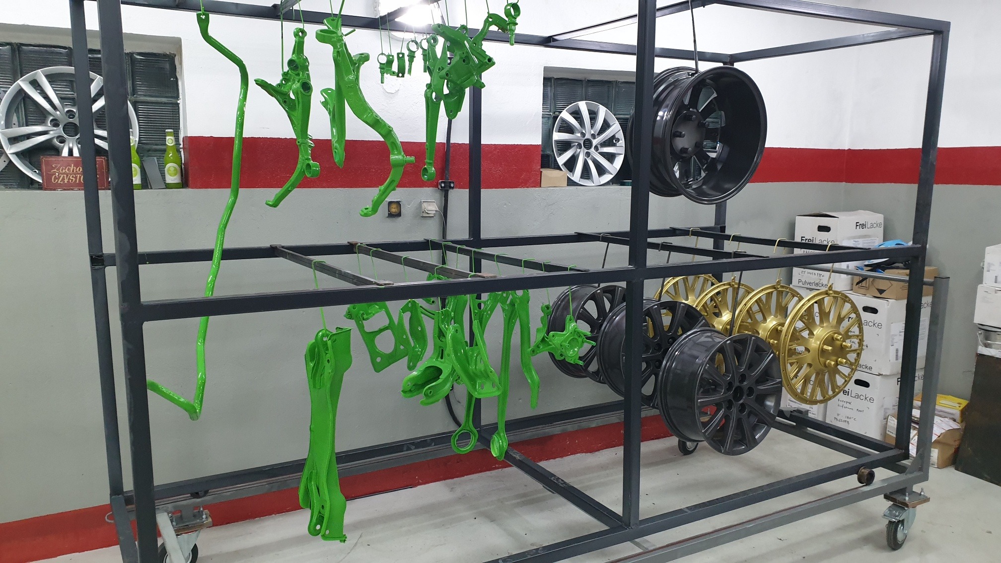 Metalowe elementy zawieszenia i felgi samochodowe po nałożeniu farby proszkowej w kolorach zielonym, czarnym i złotym, wiszące na wózku transportowym w warsztacie.