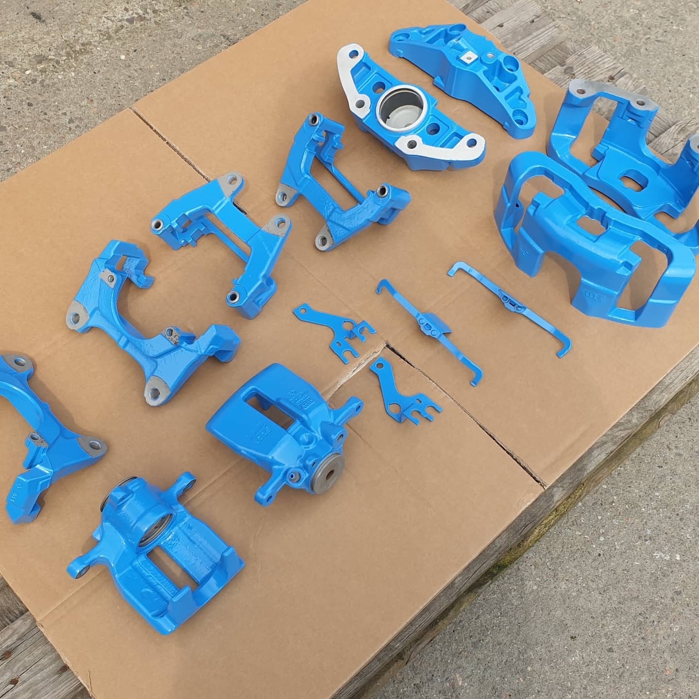 Elementy układu hamulcowego, w tym zaciski, po obróbce powierzchniowej i pomalowane na jasny niebieski kolor, leżące na kartonie.