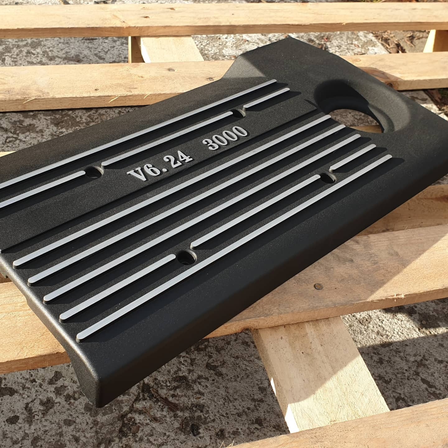 Czarna, proszkowo malowana pokrywa silnika V6.24 3000 z srebrnymi paskami, leżąca na drewnianej palecie w naturalnym świetle.