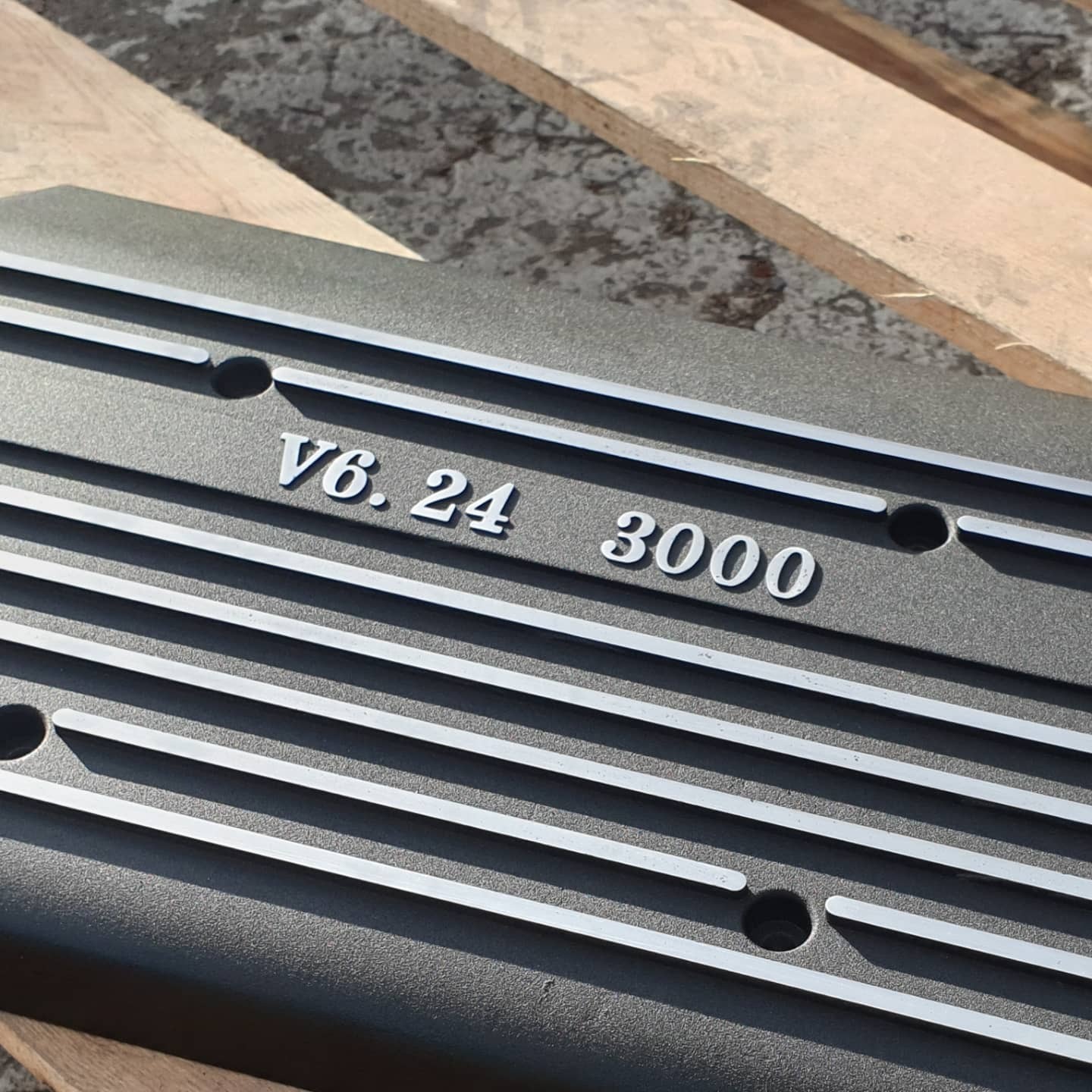 Szara, metalowa pokrywa silnika V6.24 3000 z równoległymi żebrami, leżąca na drewnianej palecie. Powierzchnia chropowata, matowa.