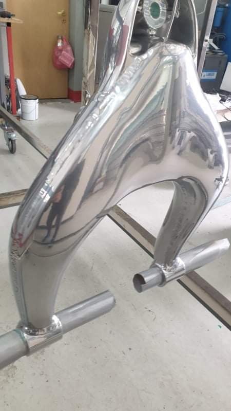 Błyszczący, metalowy element konstrukcyjny o nieregularnym kształcie, prawdopodobnie po obróbce, z widocznymi spawami i fragmentami rur, umieszczony w warsztacie.