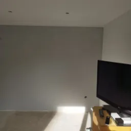 Świeżo pomalowane szarą farbą ściany w pokoju, widoczne ślady po wałku malarskim i niedokładności przy suficie, na podłodze resztki farby, w rogu telewizor na starej komodzie i wałek malarski