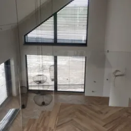 Wnętrze domu w stanie deweloperskim z widoczną drewnianą podłogą ułożoną w jodełkę, szklaną balustradą schodów, dużymi oknami z żaluzjami i wiszącymi lampami drucianymi.