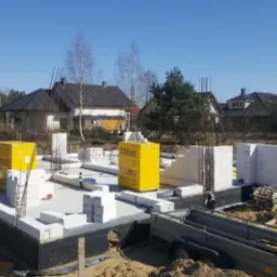 Wznoszenie ścian fundamentowych z bloczków Ytong na płycie fundamentowej, widoczne zbrojenia i przyczepka transportowa na placu budowy w słoneczny dzień.